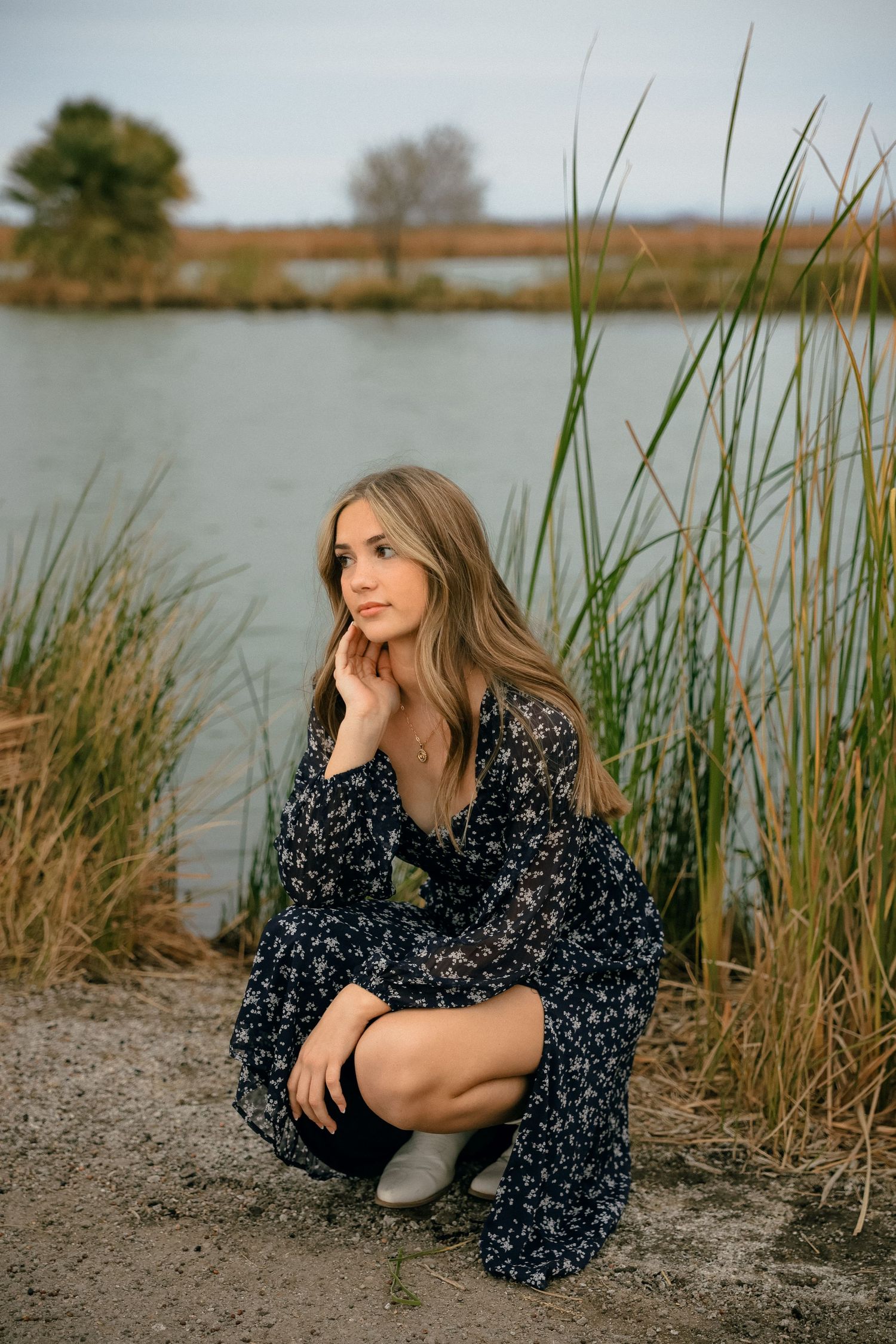 A woman in a floral dress crouches by a lake surrounded by tall reeds and grasses at golden hour.