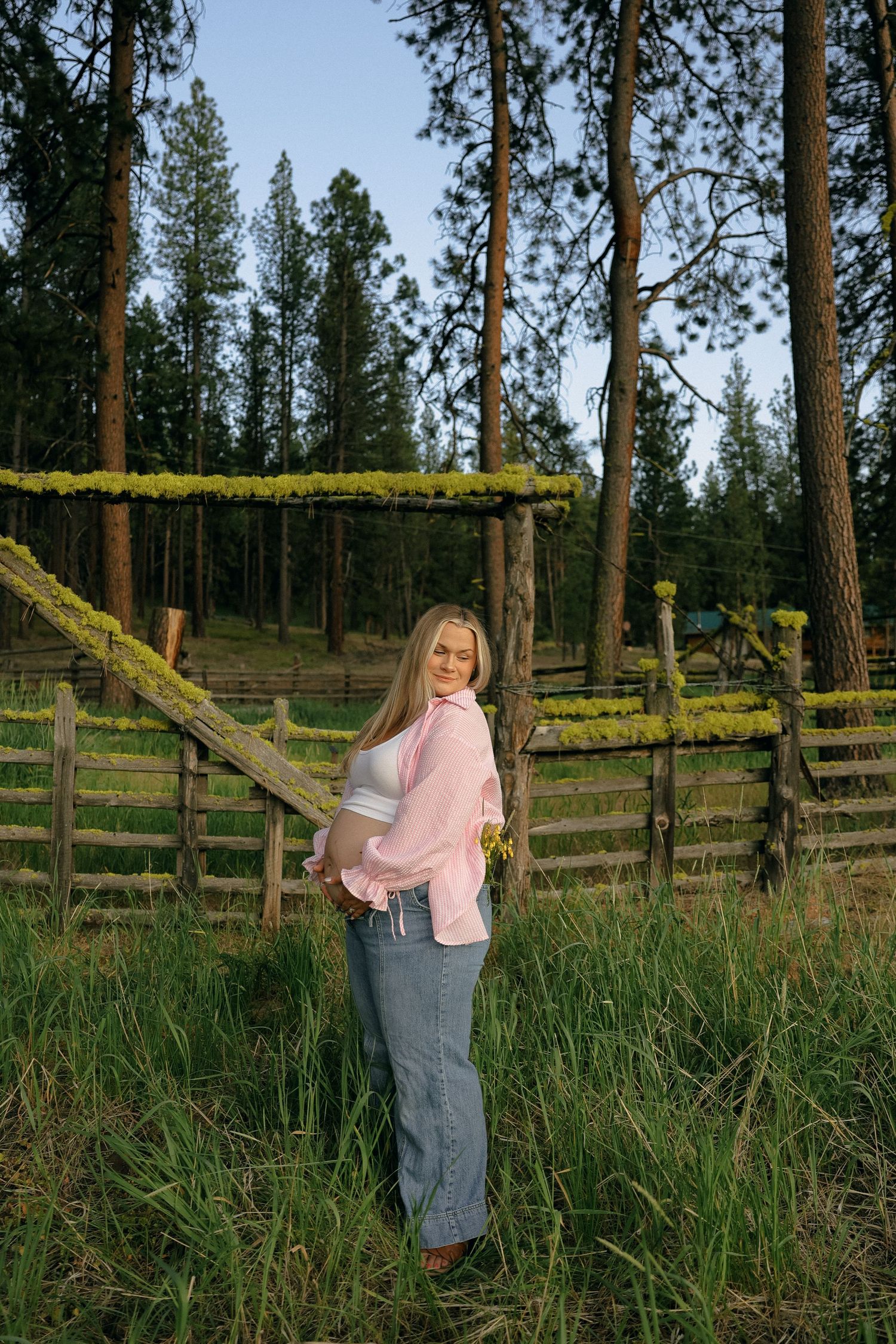 Someone in a pink sweater and denim skirt stands by a rustic wooden fence surrounded by tall pines and grass.