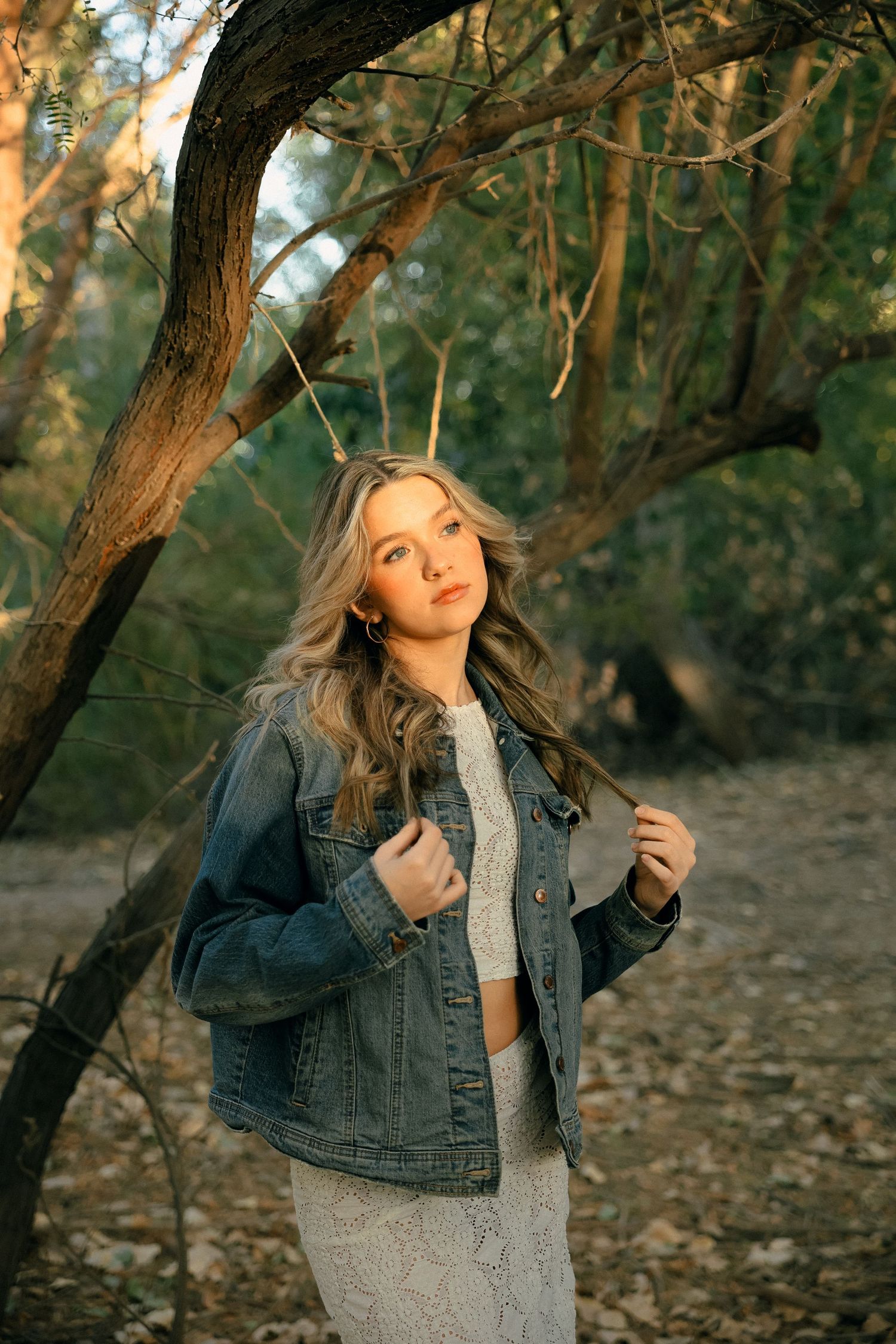 A woman in denim jacket poses against tree branches in warm afternoon sunlight.