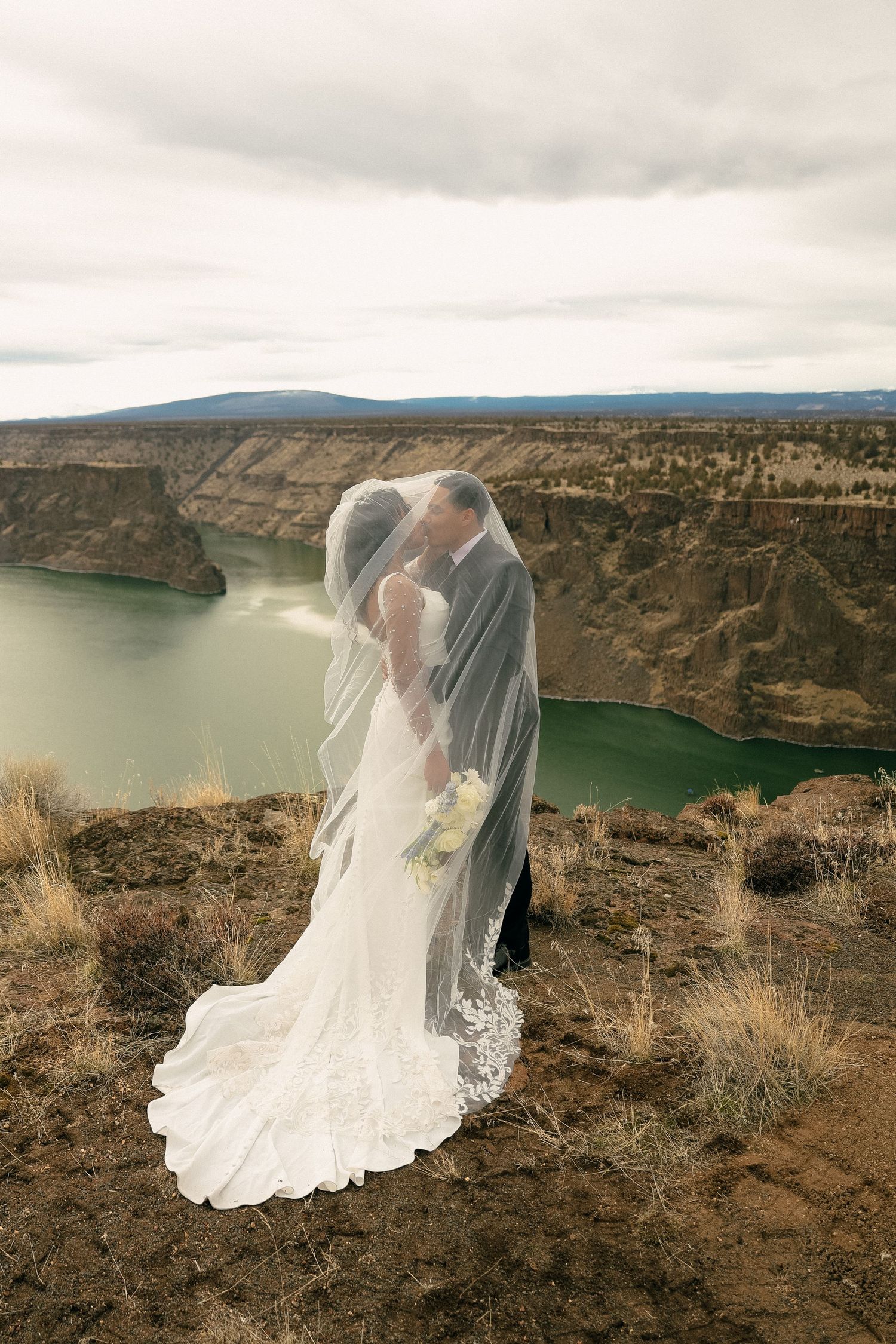 Wedding couple standing on cliff edge overlooking scenic lake at sunset.