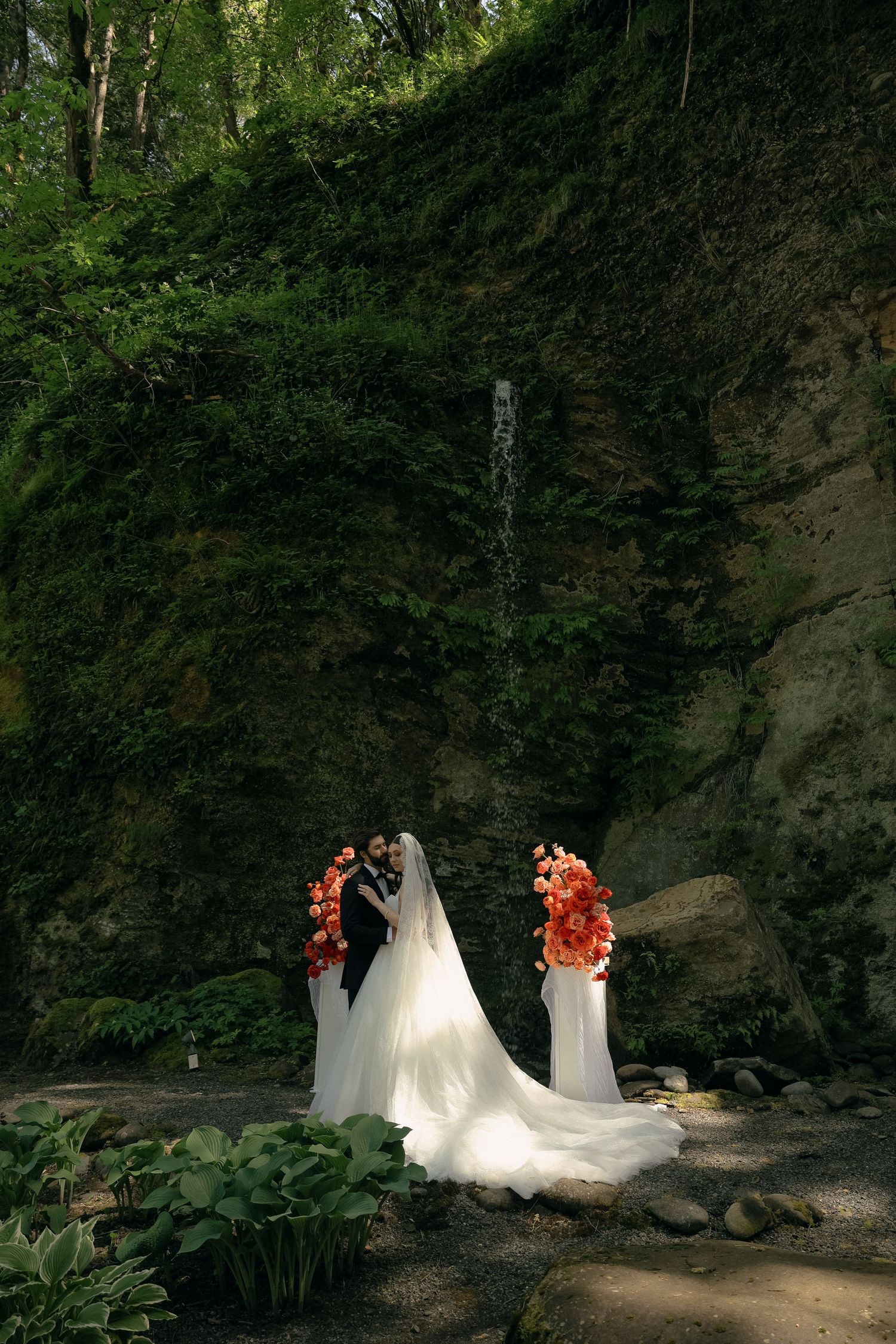 Dramatic wedding dress train displayed near natural waterfall with coral floral arrangements.
