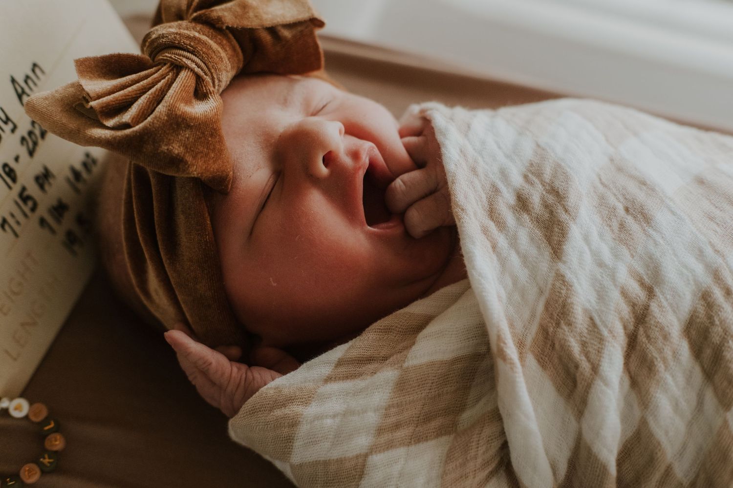 A baby wearing a brown headband yawns while laying on cream colored textured bedding.