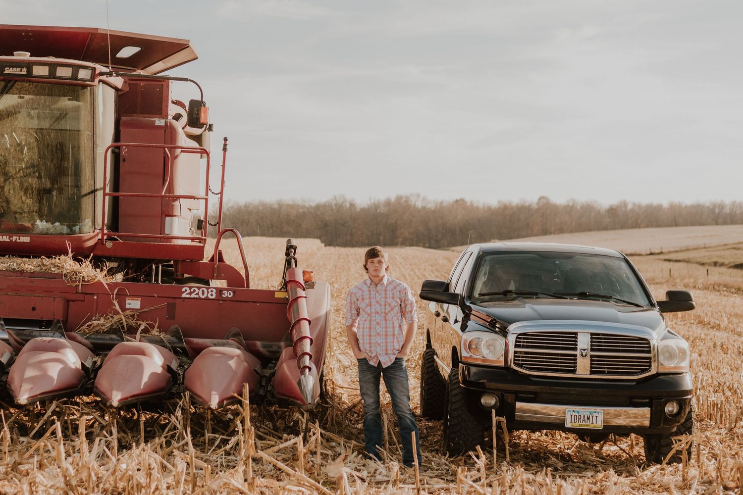 A senior boy poses by their truck and red combine harvester in a rural farm field.
