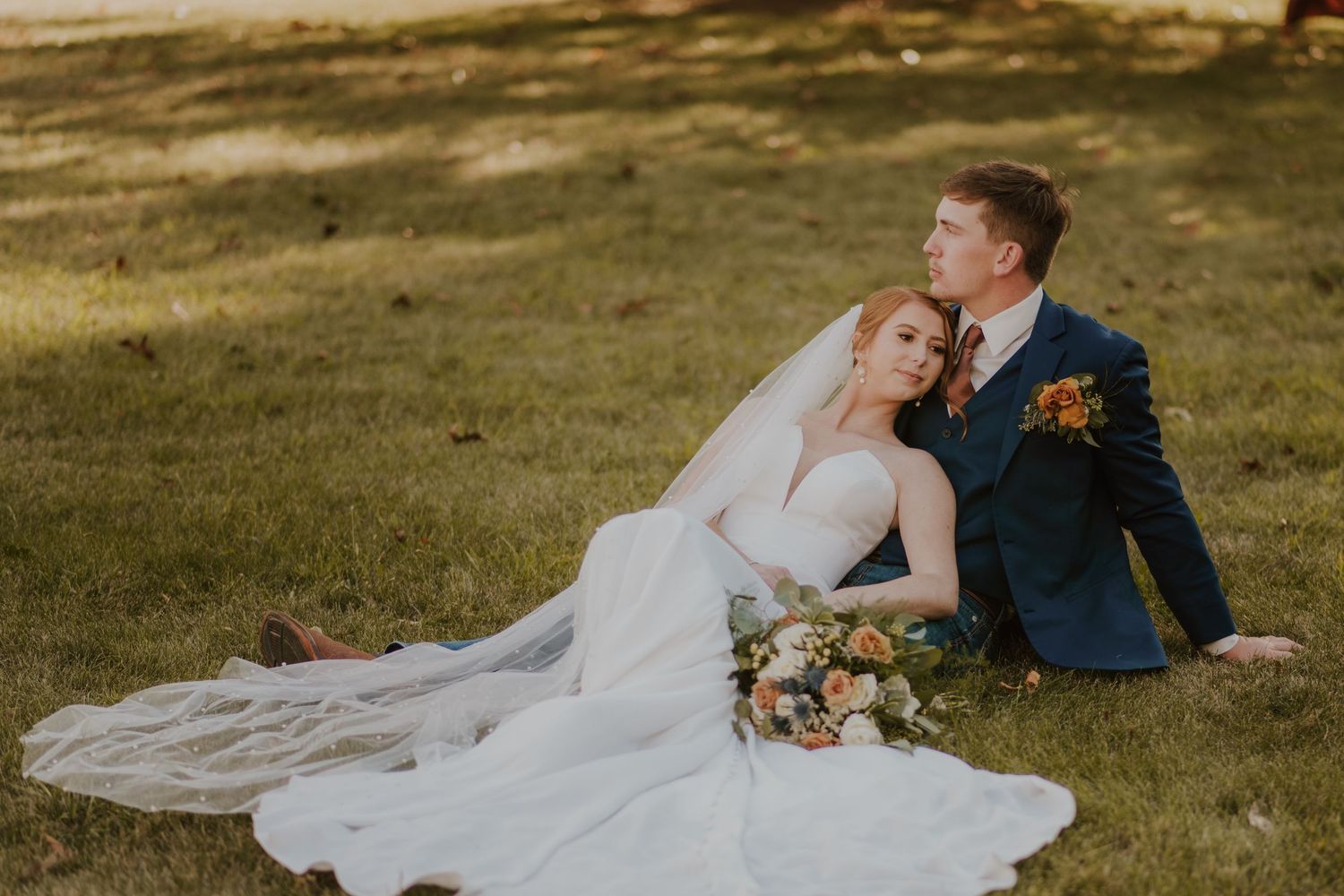 A bride in white gown and groom in navy suit sit together on grass with autumn wedding bouquet at sunset.