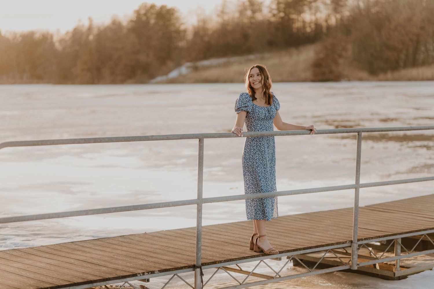 A senior girl in a blue dress stands on a wooden dock during sunset.