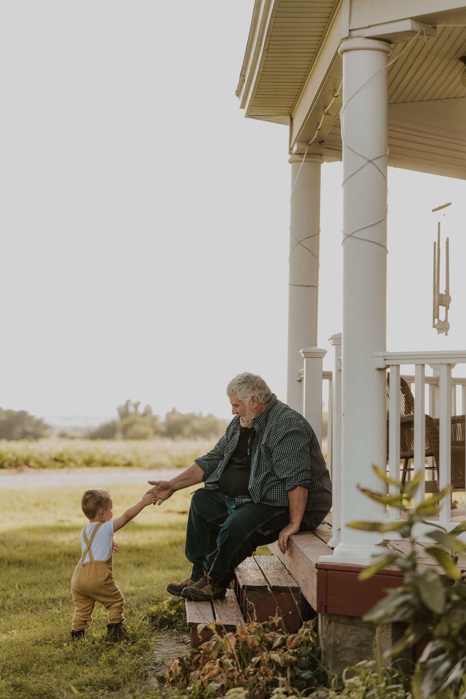 Sweet moment captured on a farmhouse porch showing an elderly person and small child holding hands in golden sunlight.