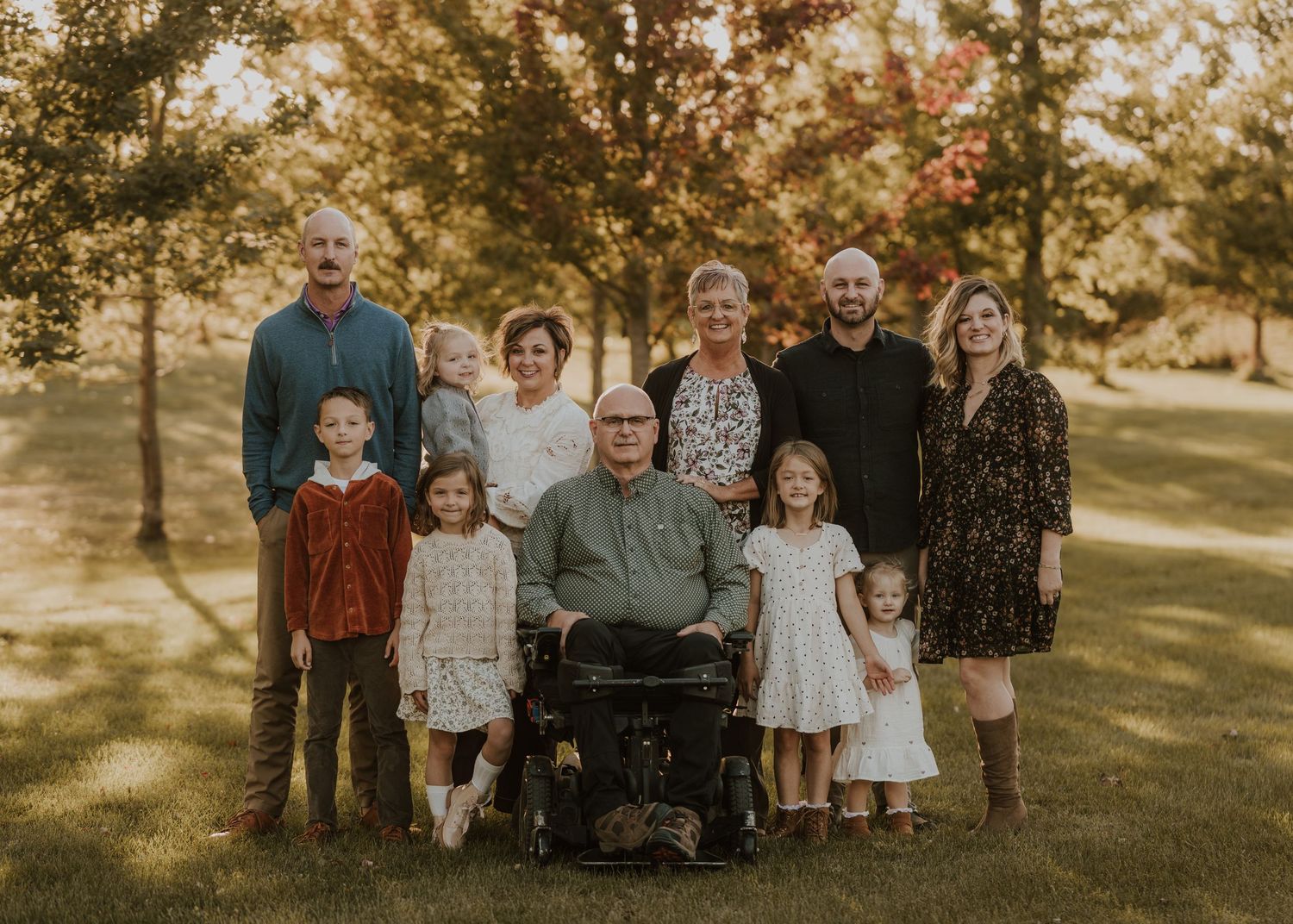 A multi-generational family poses together outdoors in a park during golden hour with autumn trees in the background.