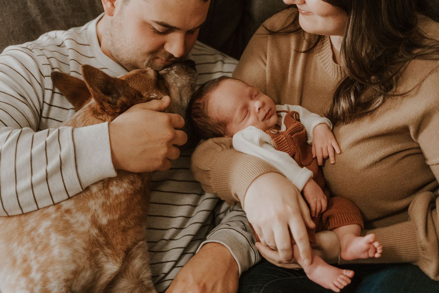 A couple holds their newborn baby while sitting on a couch with their red Healer dog close by.