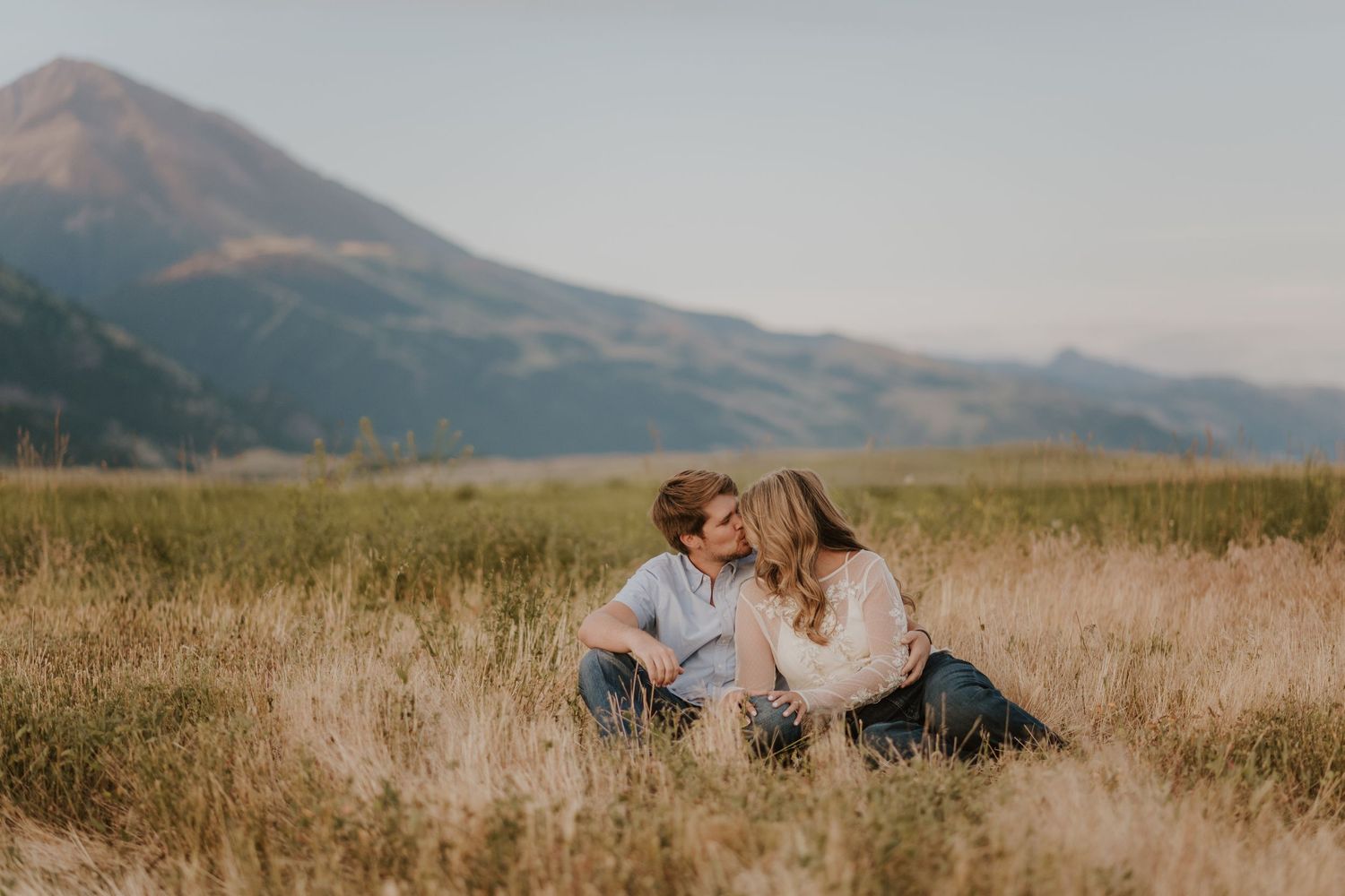 A couple sits in tall grass with mountains in the background during sunset.