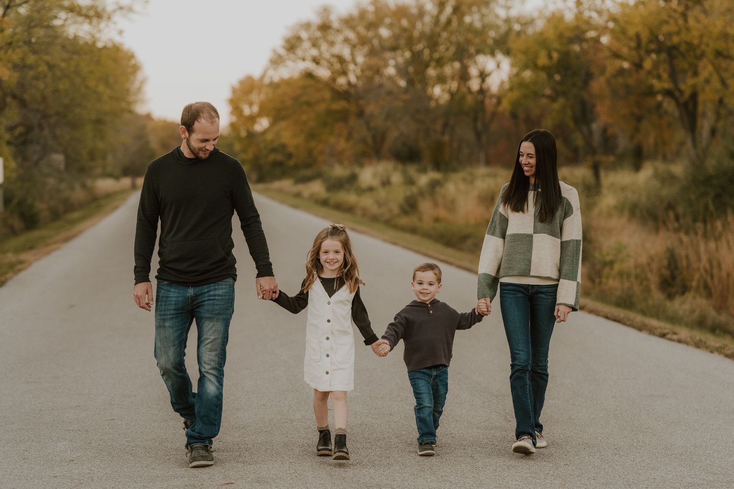 A family of four walks hand in hand down a rural road during autumn.