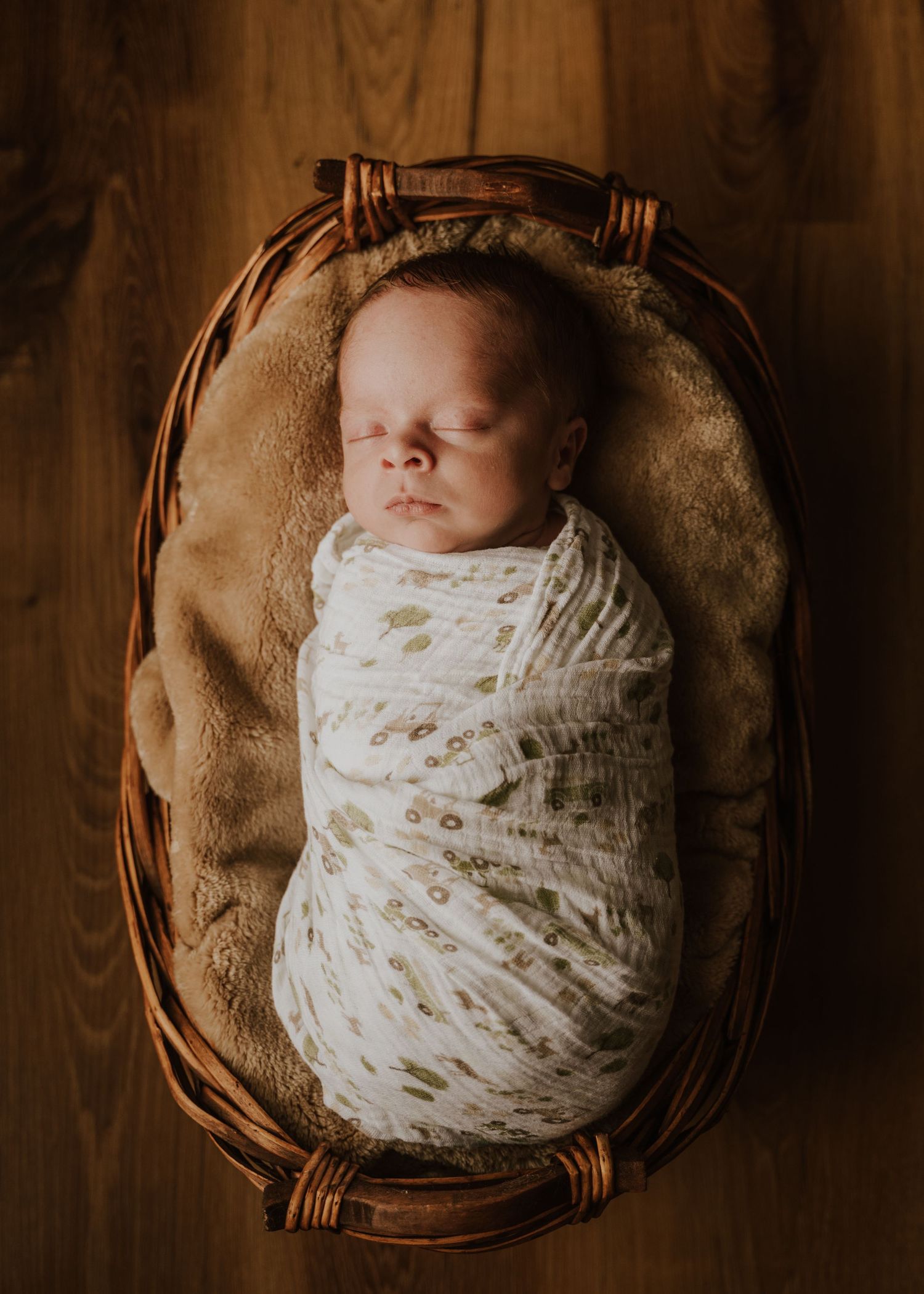 A sleeping newborn wrapped in white muslin swaddle lies peacefully in a brown wicker basket.