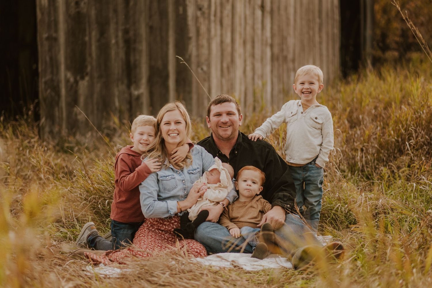 A family sits together in tall autumn grass near a rustic wooden barn on a warm golden afternoon.