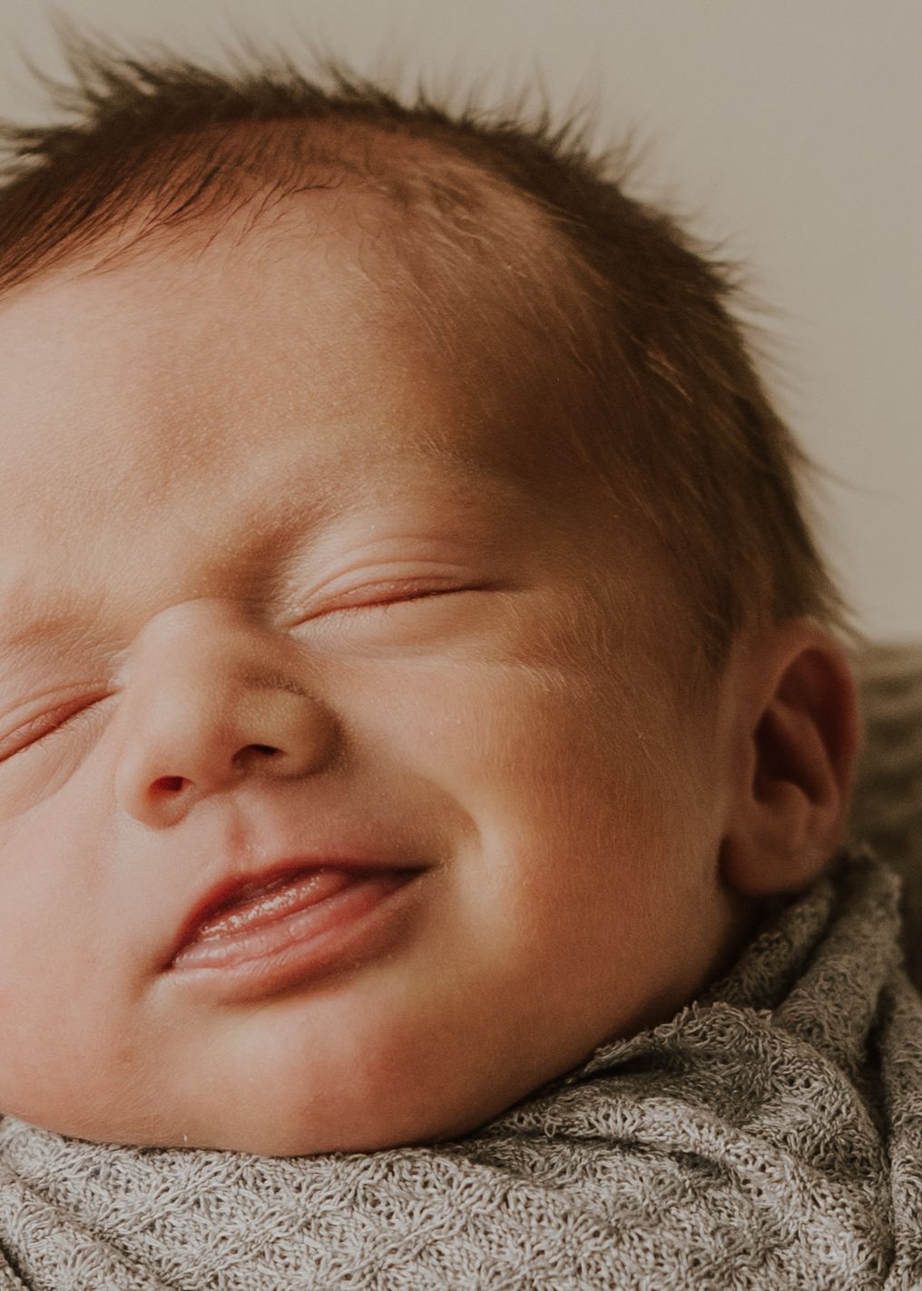 Close up of a smiling baby with closed eyes and rosy cheeks against a neutral background.