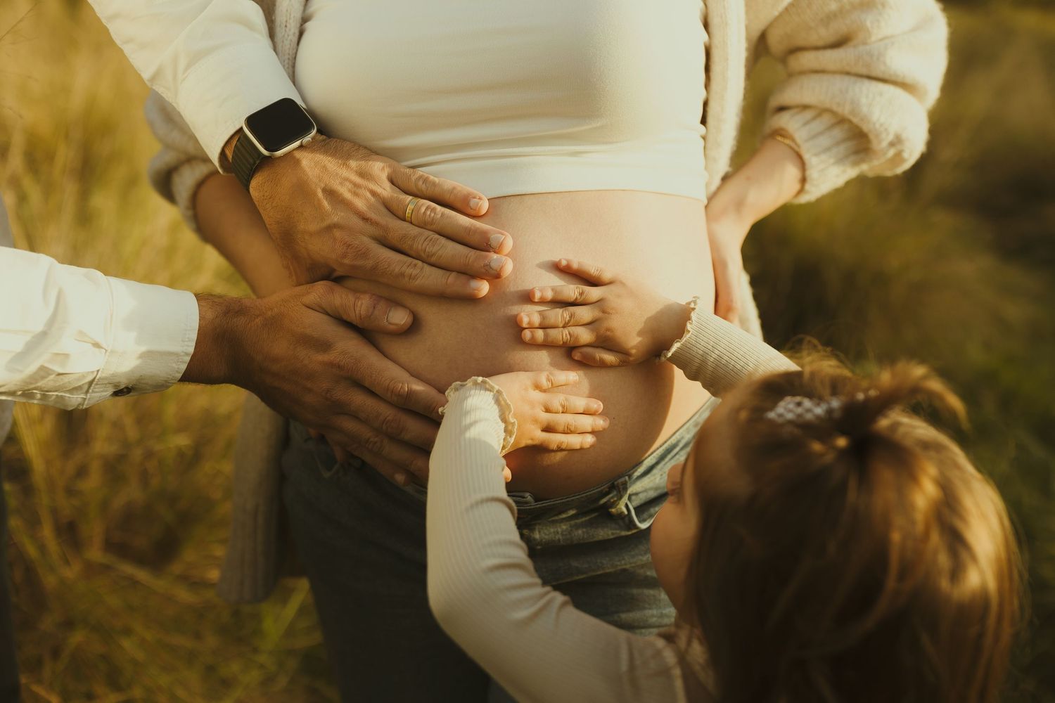 Close-up shot of hands touching pregnant belly in warm sunset lighting outdoors.
