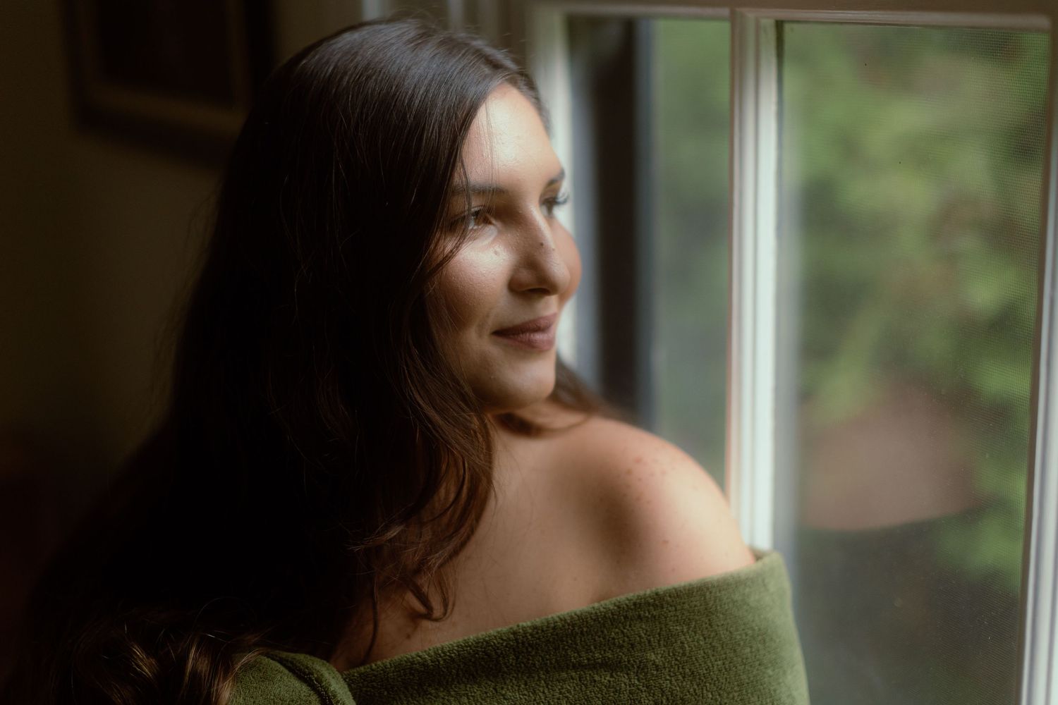 Soft portrait by window with dark long hair and olive sweater gazing in natural light.