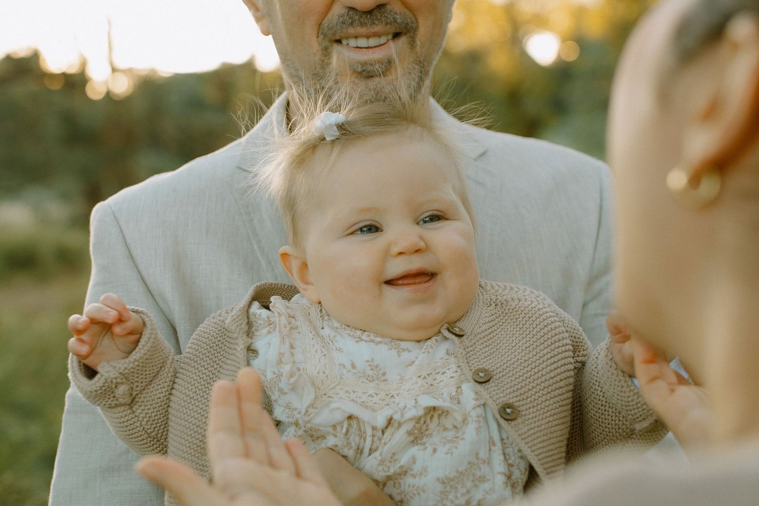 A baby smiles joyfully while wearing a white lace dress and sitting outdoors during a sunny summer day.