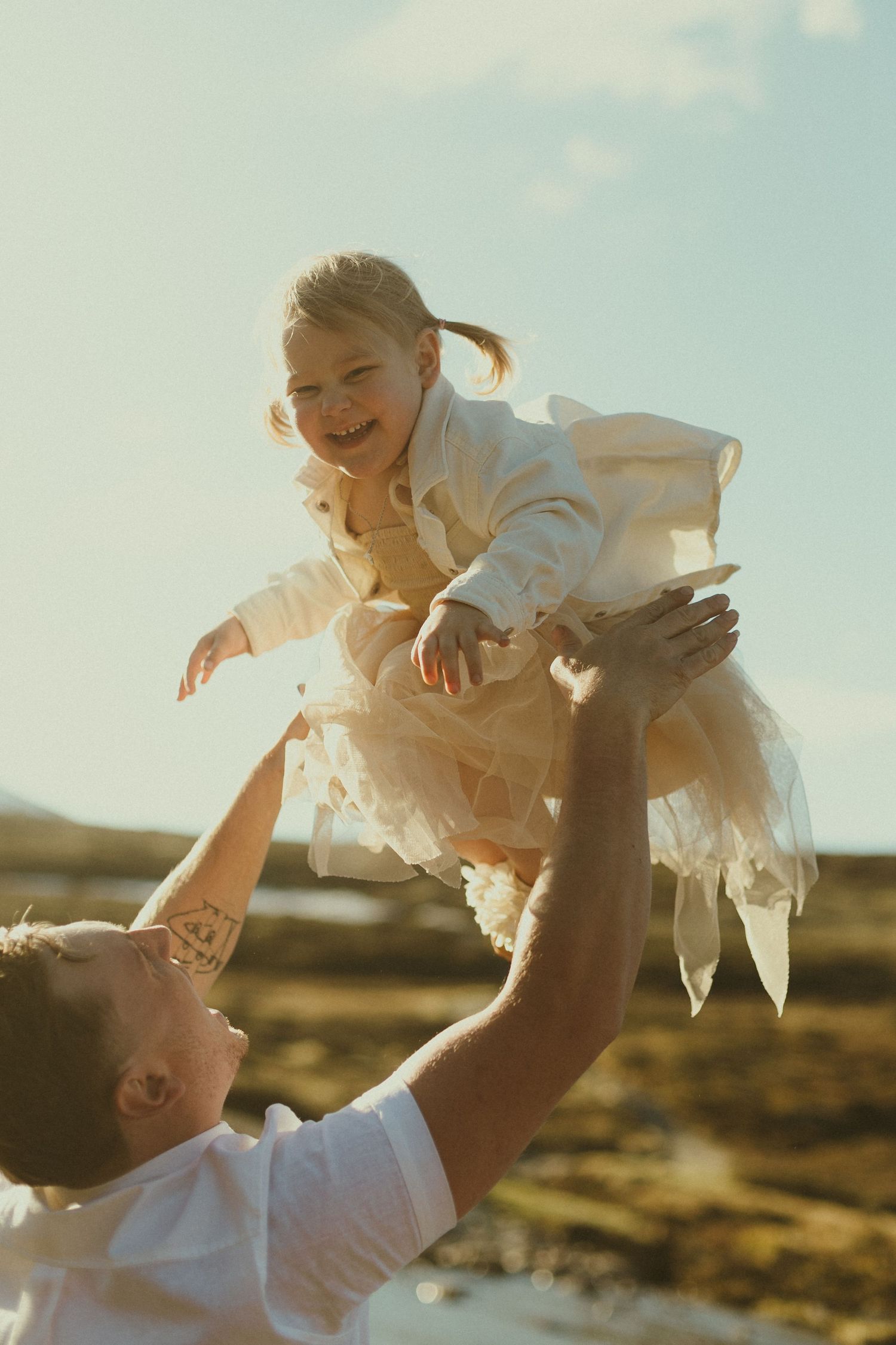 A joyful moment captured at sunset as a person lifts a child wearing a white dress into the air against a golden sky.