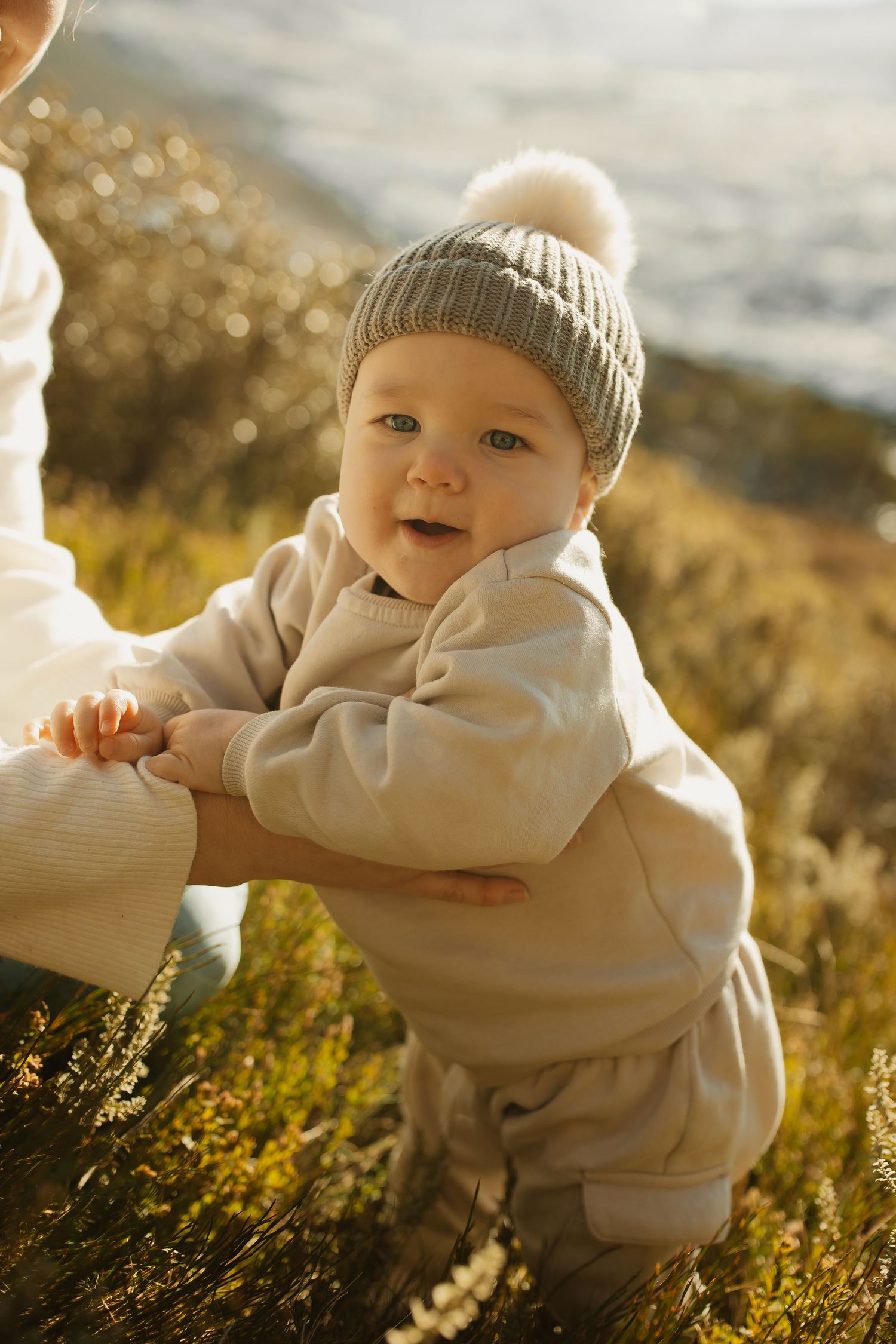 Small child wearing a gray winter hat and beige outfit stands in sunlit field during golden hour.