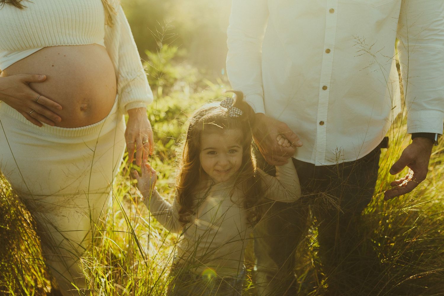 Golden hour family photo shoot in tall grass with expectant mother and child sharing a tender moment.