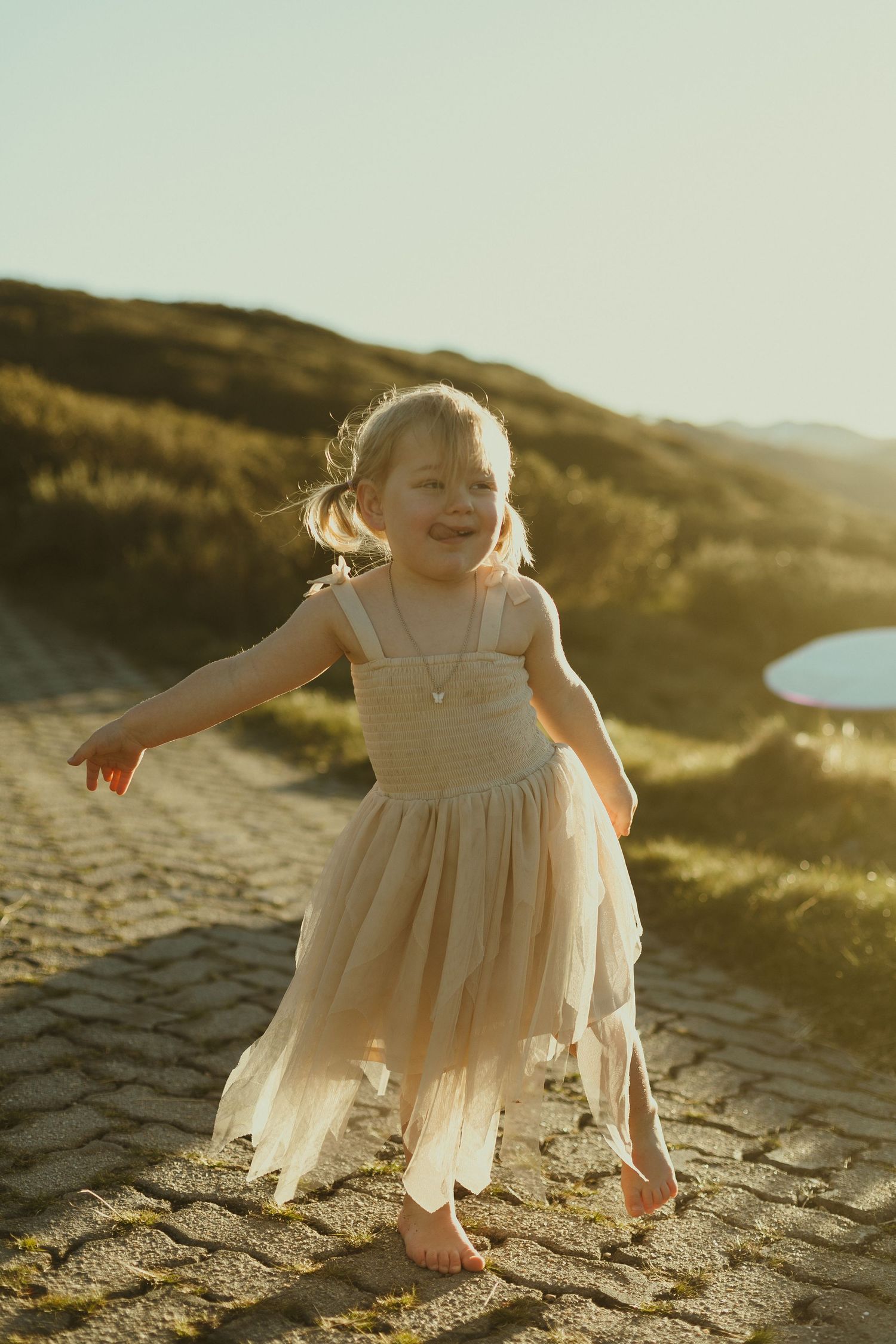 Young child wearing a flowing cream dress dances joyfully on a stone path during golden hour.