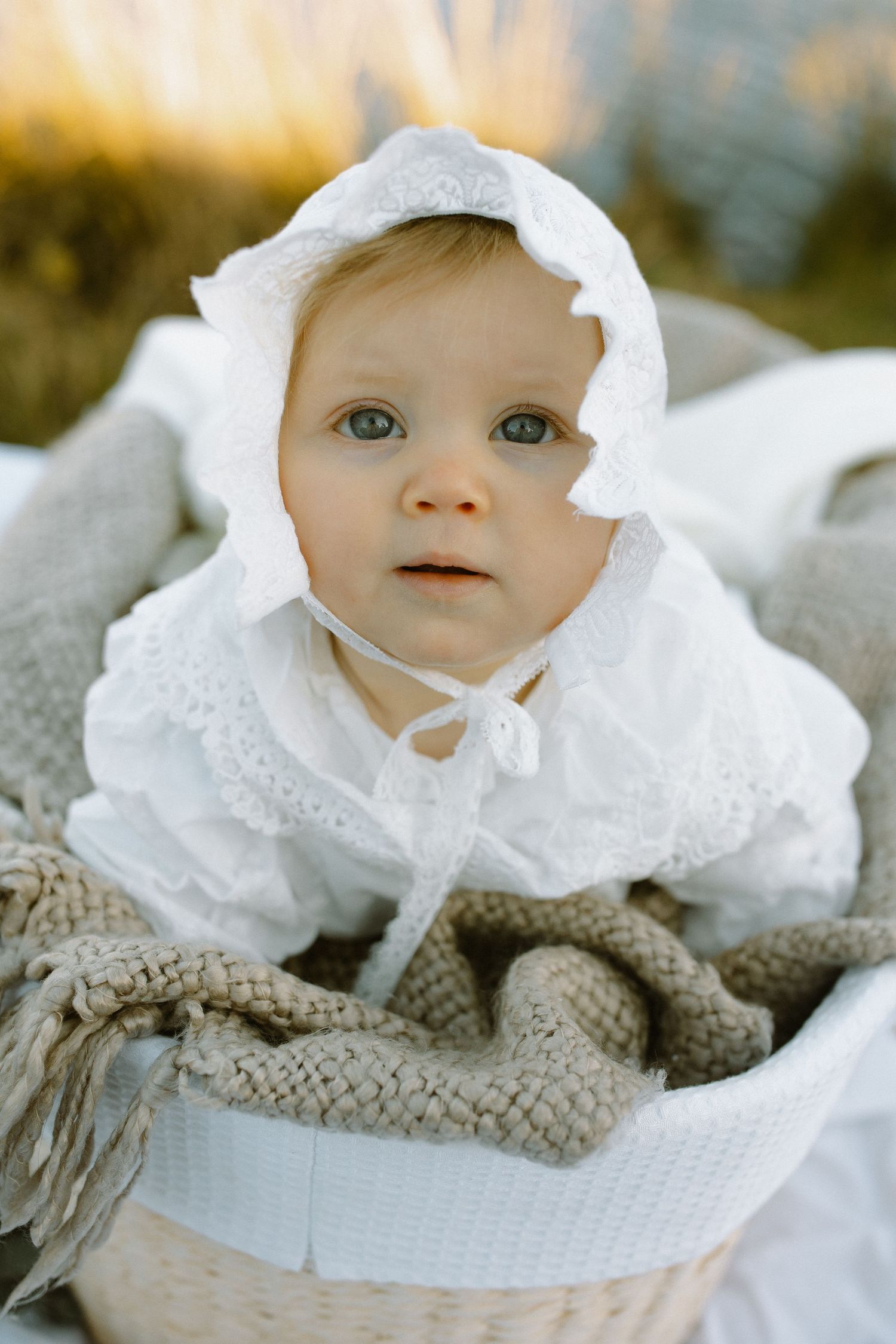A small child wearing a vintage-style white christening gown and bonnet rests on a knitted blanket in soft lighting.