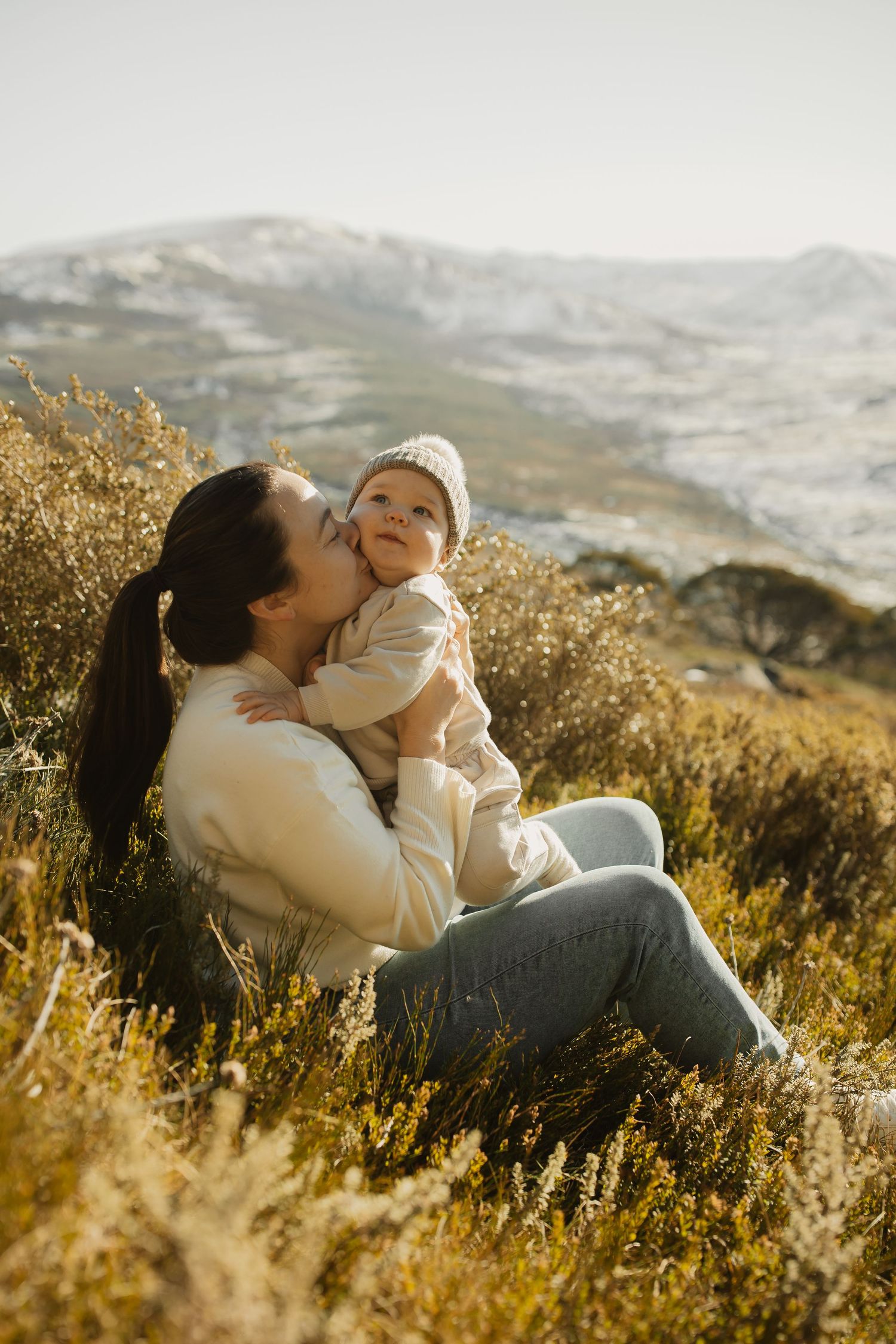 A tender moment shared between two people sitting in golden mountain grass at sunset with snow-capped peaks in the distance.