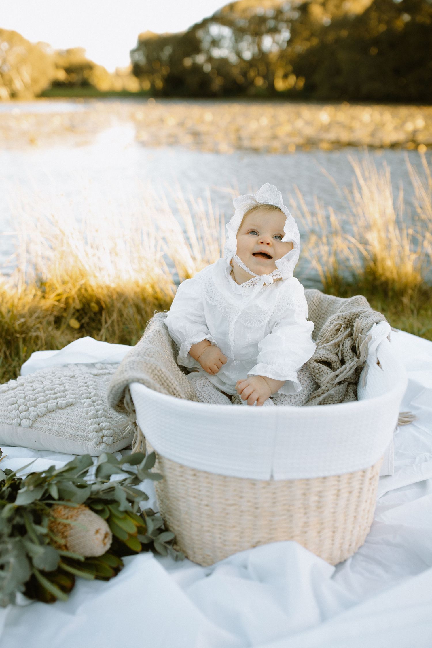 A baby wearing white sits in a woven laundry basket on a blanket by a lake during golden hour.