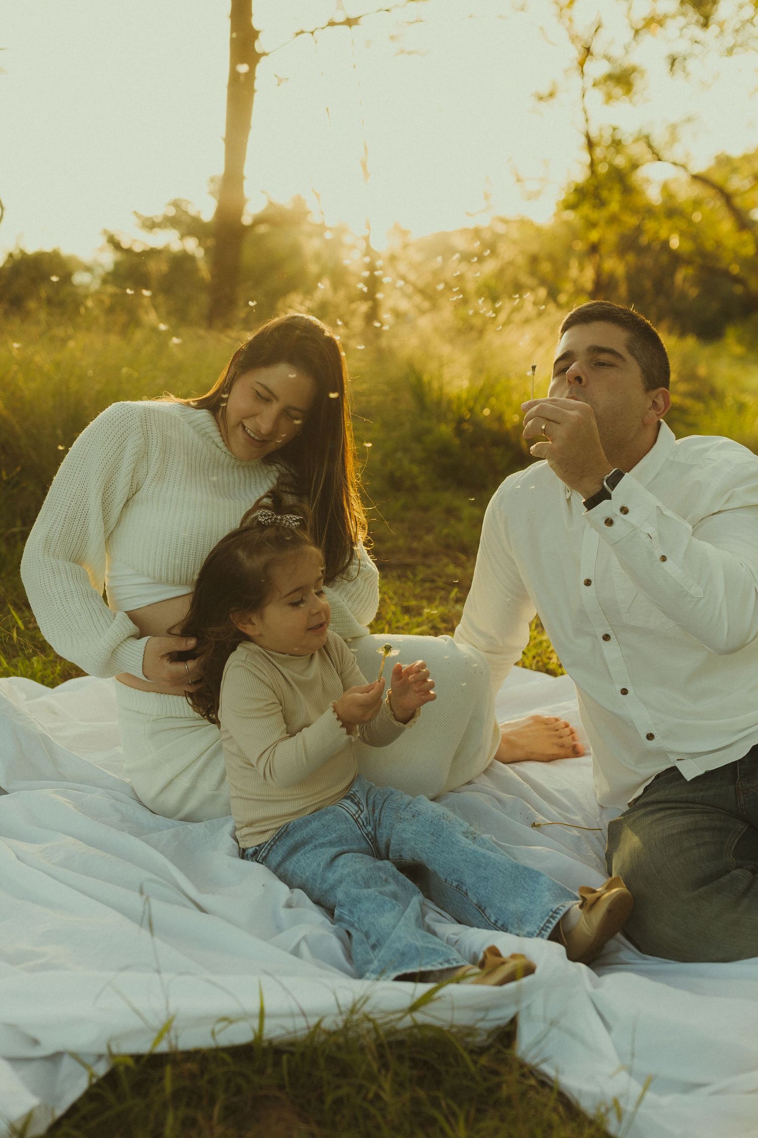 Family sitting on white blanket in sunlit field during golden hour sharing tender moment together.
