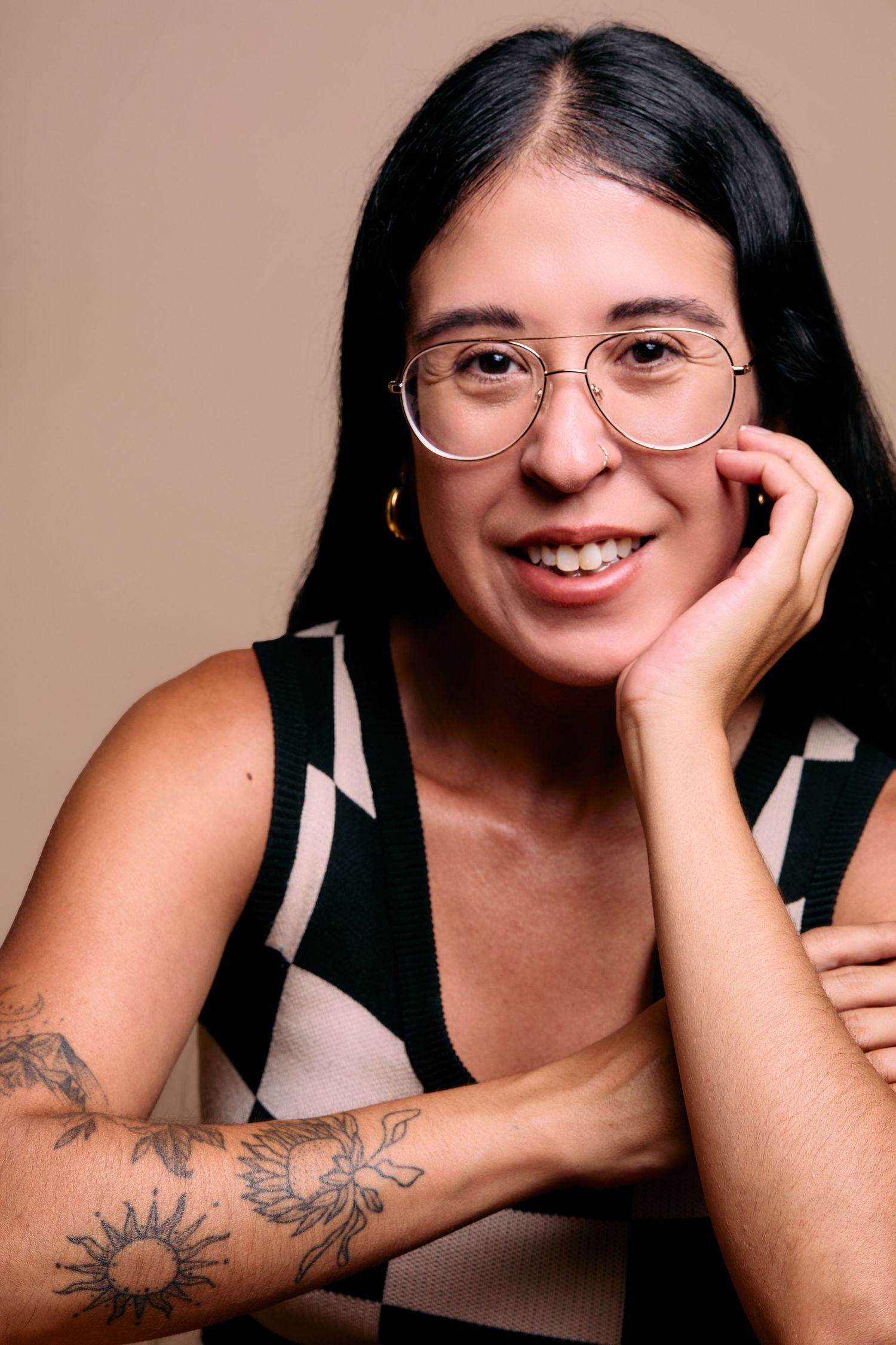 Portrait against pink background showing round glasses, arm tattoos and casual black top with geometric pattern details.
