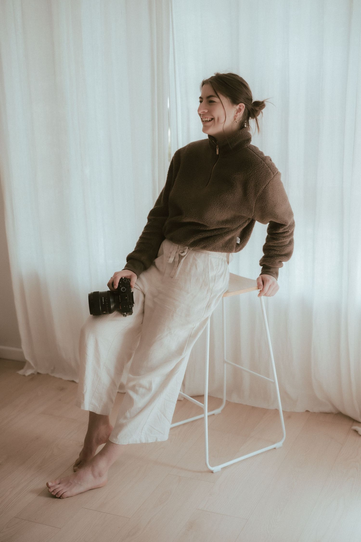 Person in brown sweater and white pants sits casually on stool holding camera.