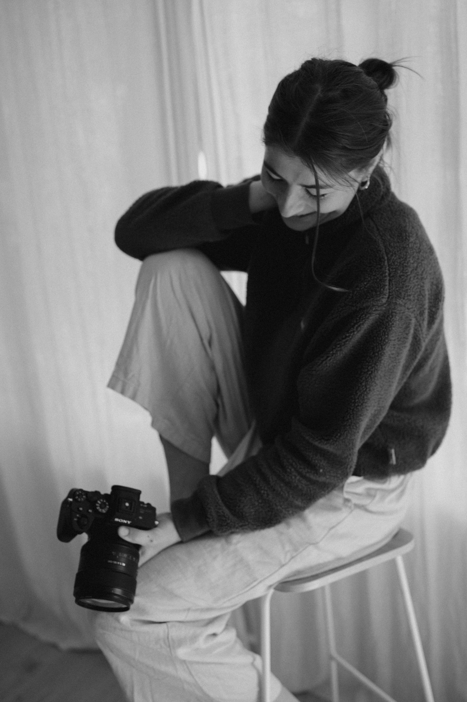 Photographer in cozy sweater sits on white stool with camera in monochrome setting.
