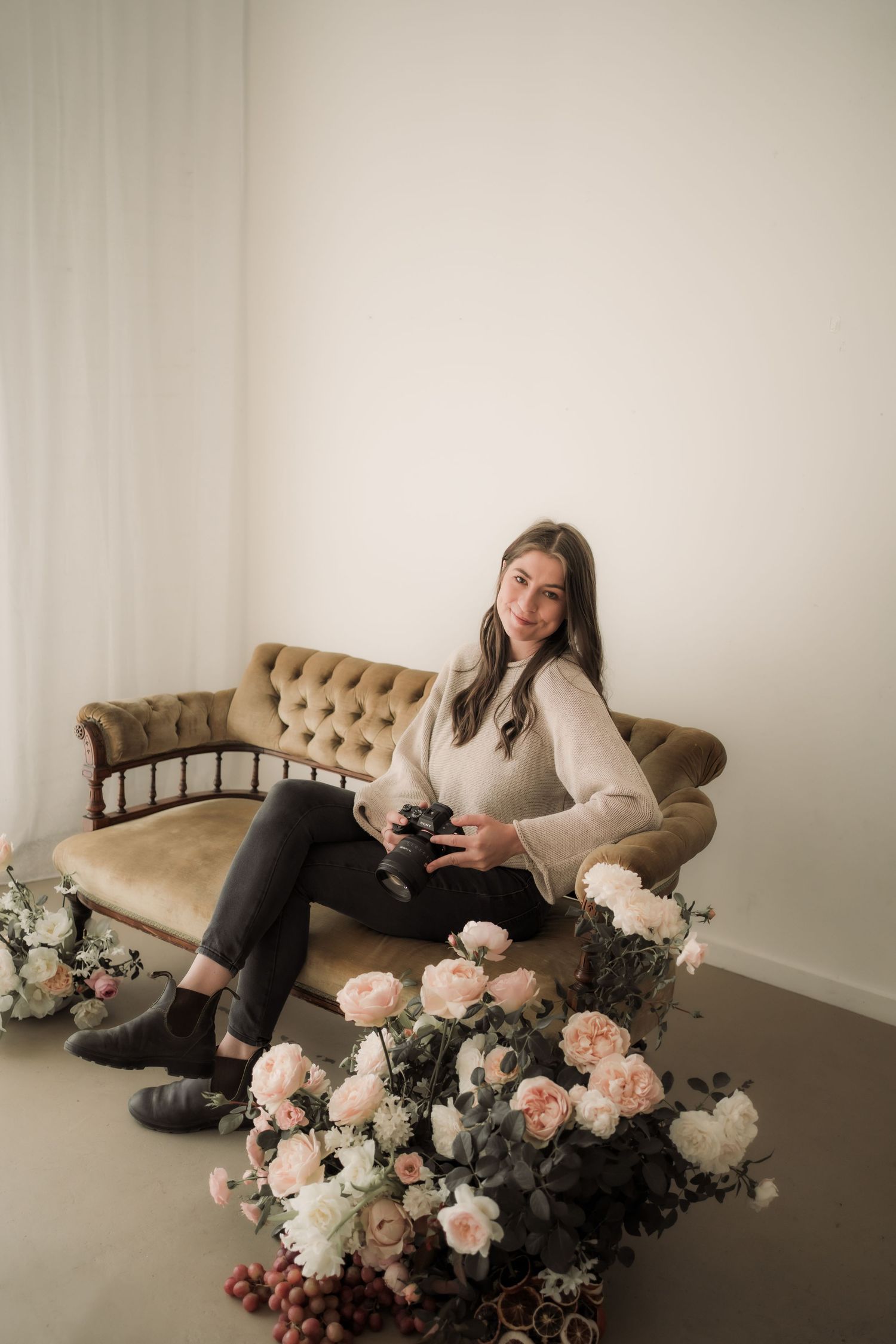 Cozy lifestyle scene with person relaxing on tufted beige sofa surrounded by lush pink and white floral arrangements.