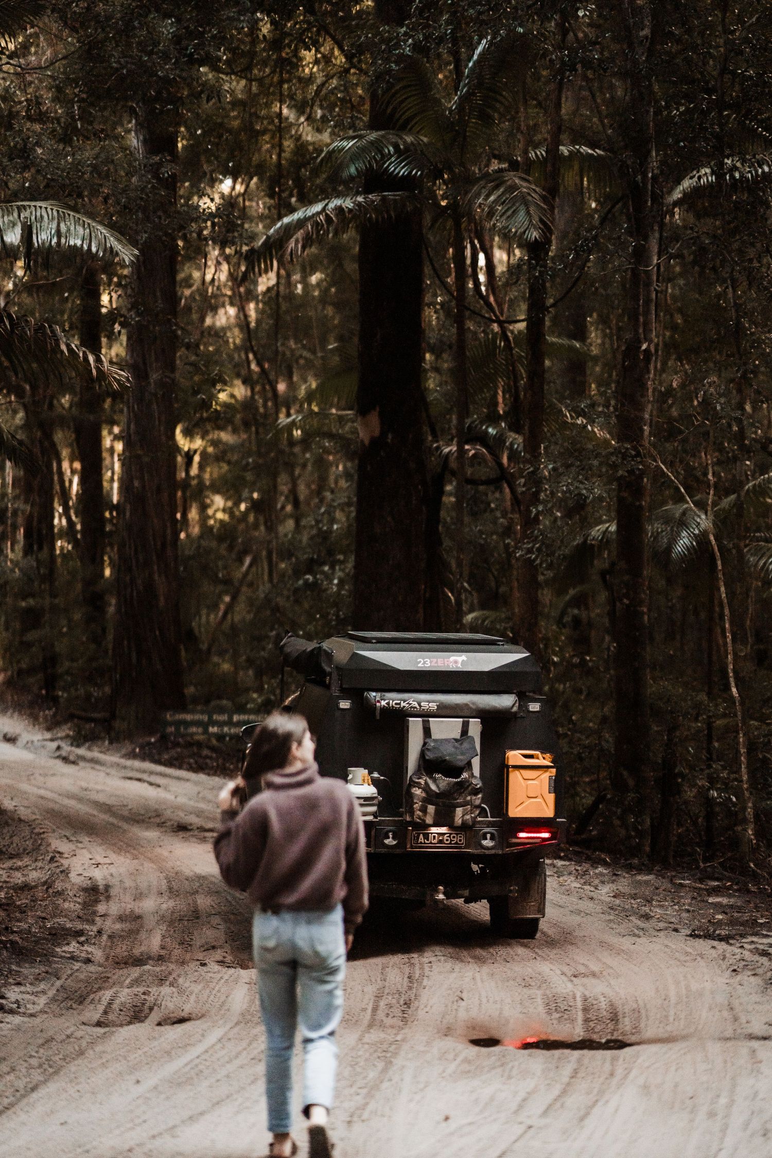 Person walking down a dirt road in a forest following a vehicle with a rooftop tent setup at dusk at Fraser Island, K'gari