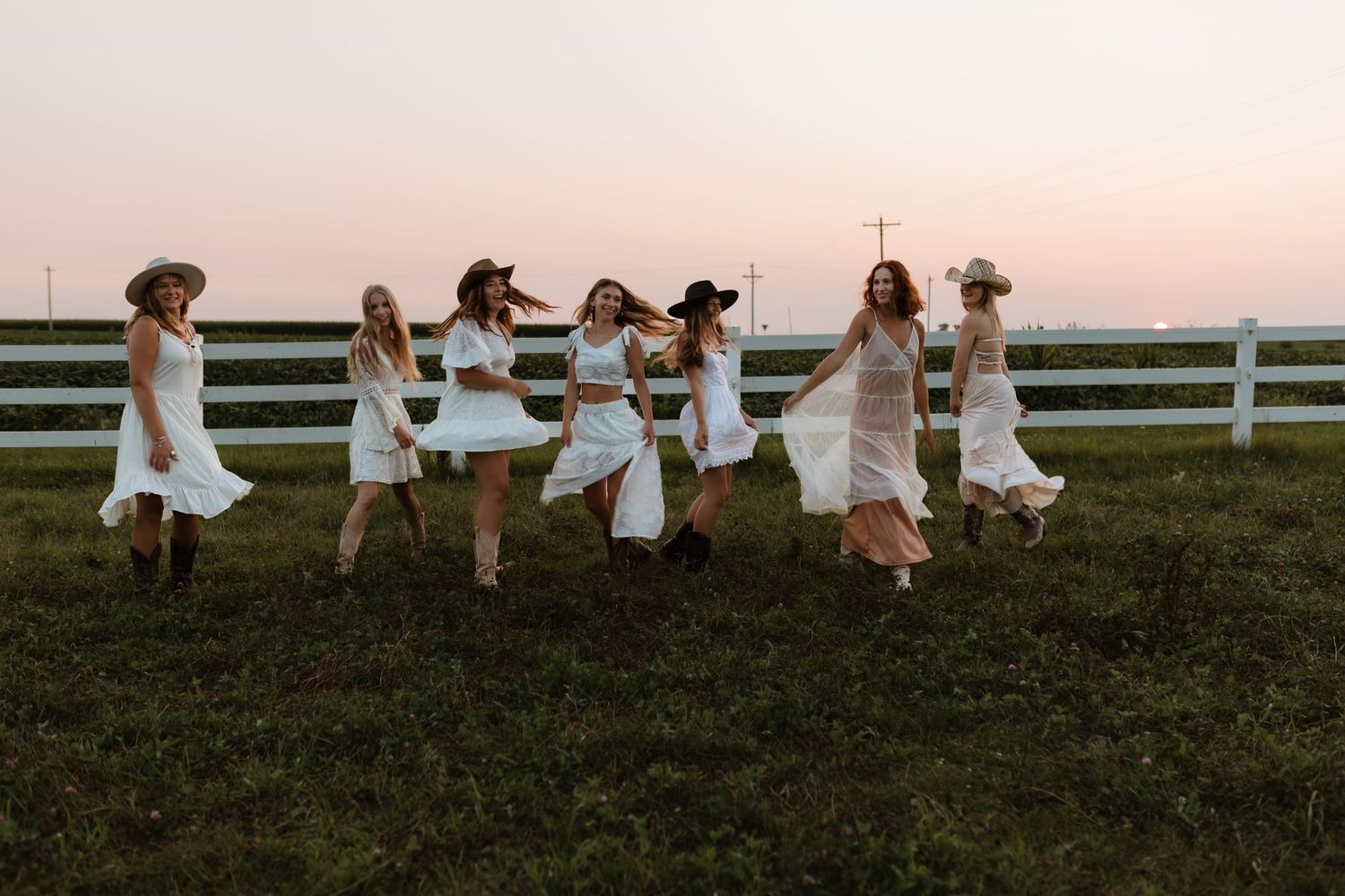 A group of women in white dresses jumping together in a field at sunset.