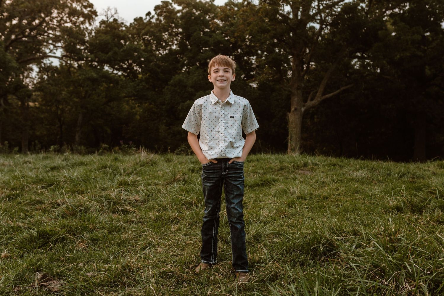 Boy in button-down shirt poses in grassy field during sunset.