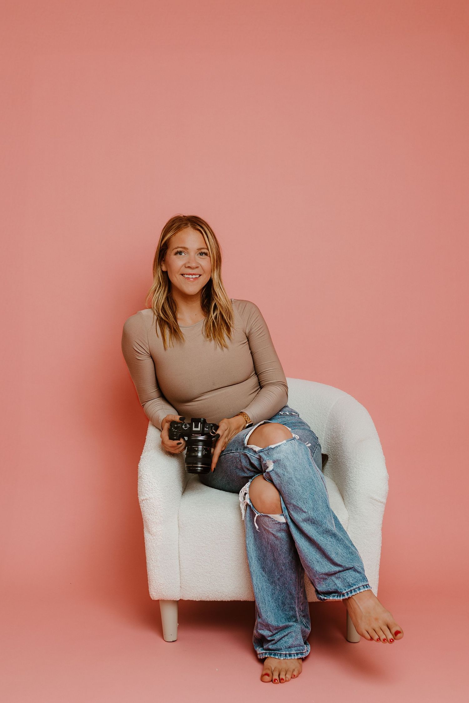 Casual portrait of photographer sitting in white chair with camera against coral background.