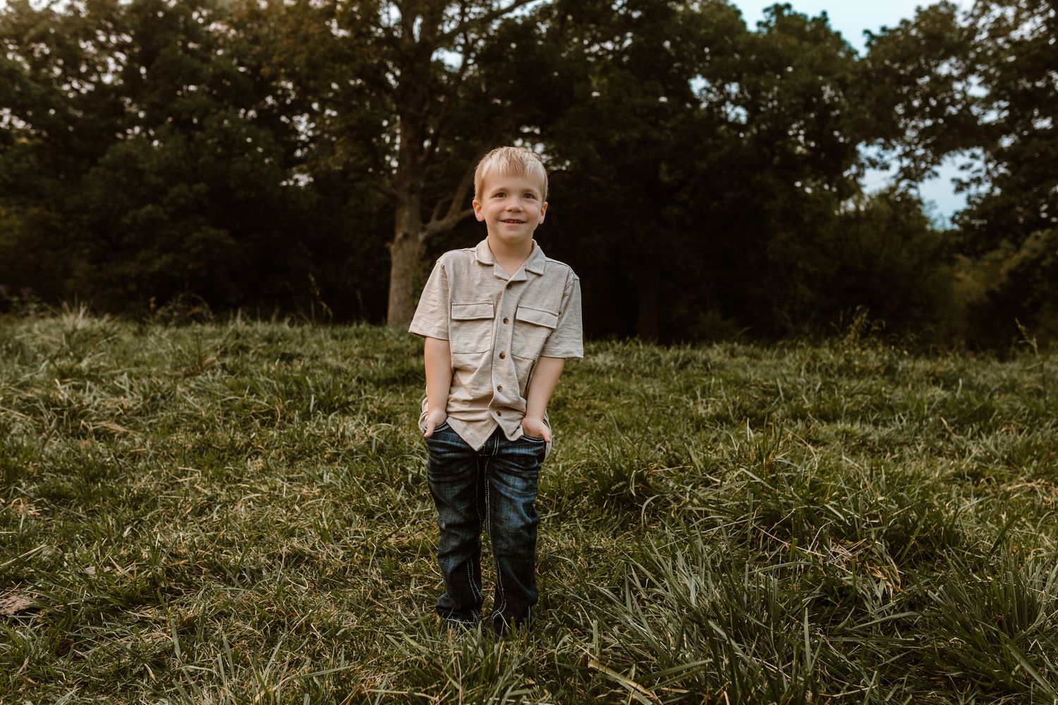 Young figure wearing a tan shirt poses in a green field with trees in the background.
