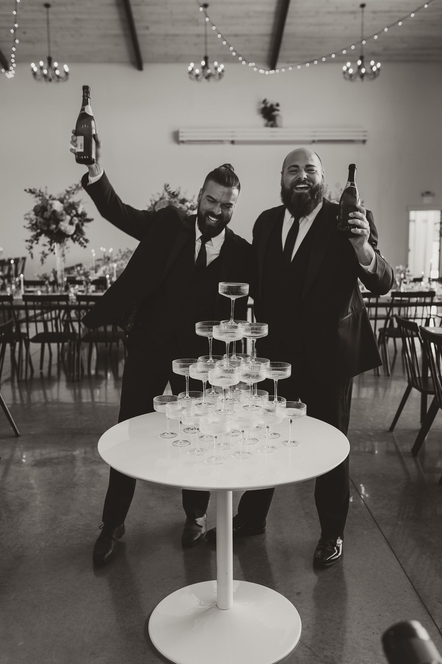 Two men in suits celebrating with champagne tower at wedding reception.