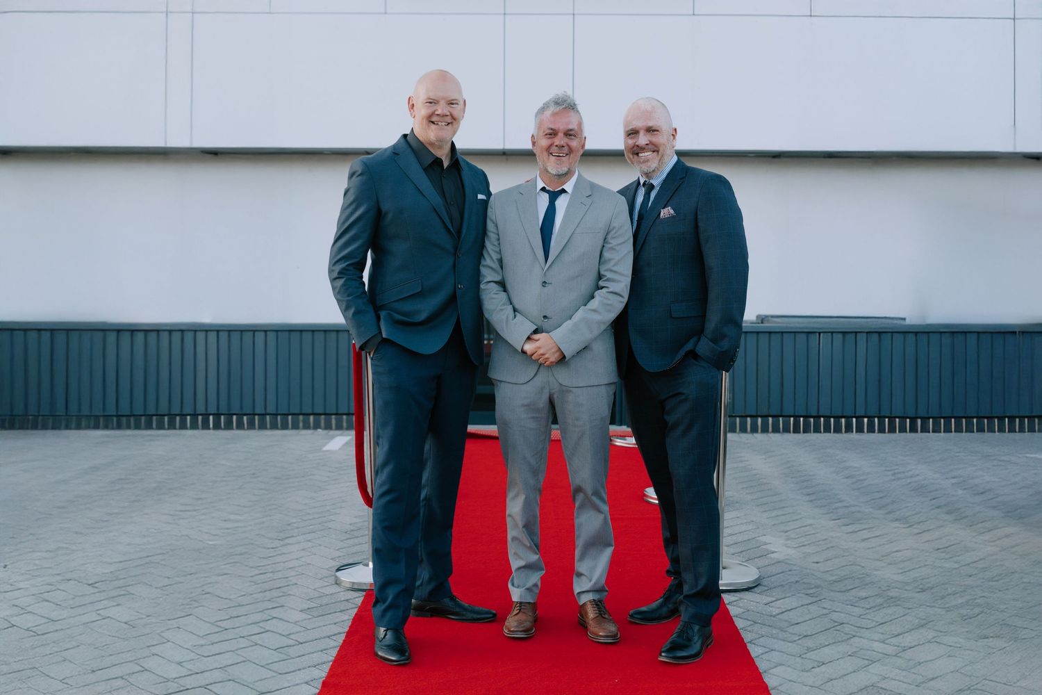 Three men in suits stand on a red carpet outdoors in front of a modern building at a corporate gala event photography in Basildon Essex
