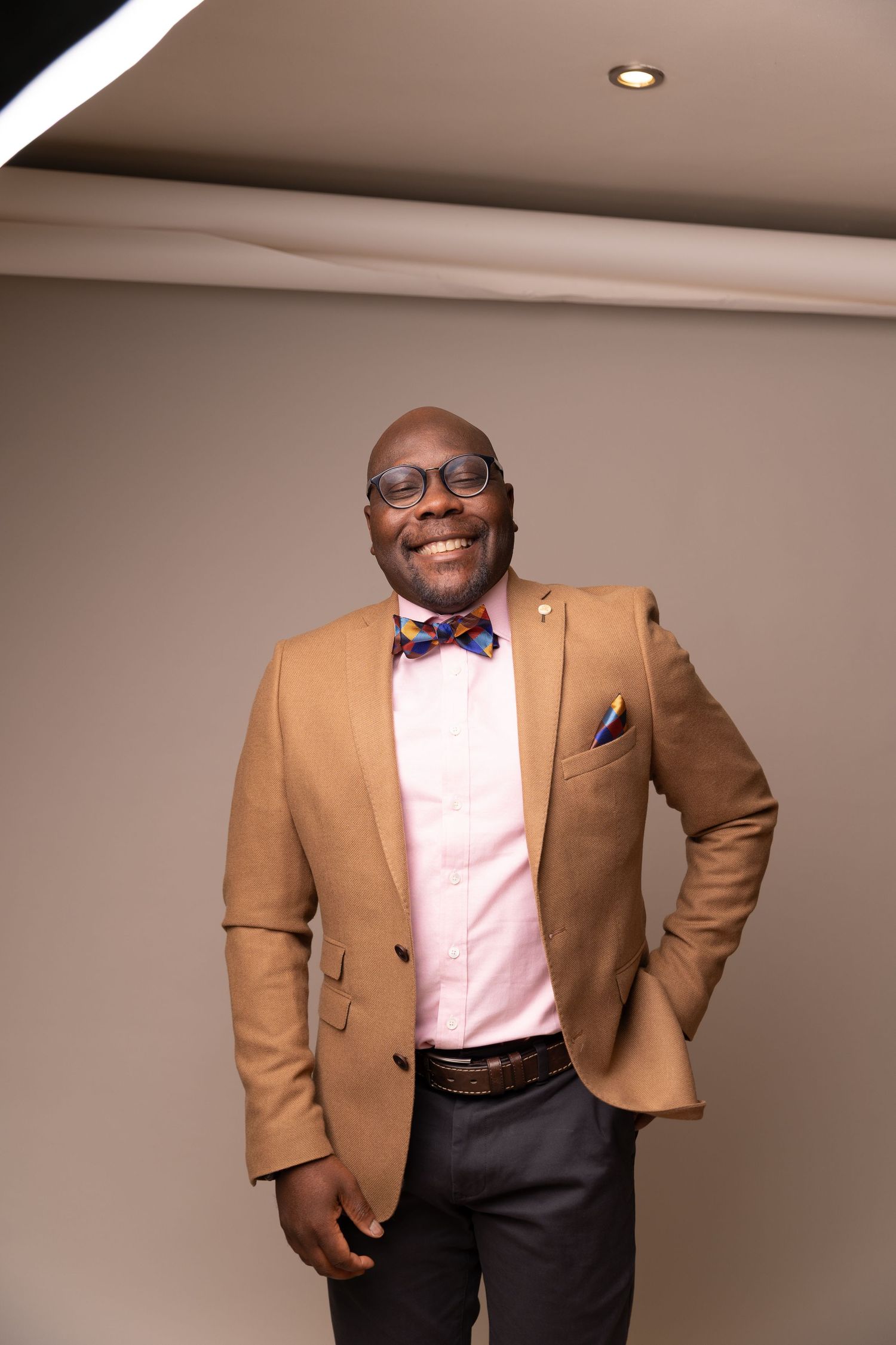 unedited headshot of a smiling man in glasses, a tan blazer and colorful bow tie, and dark pants at photo studio