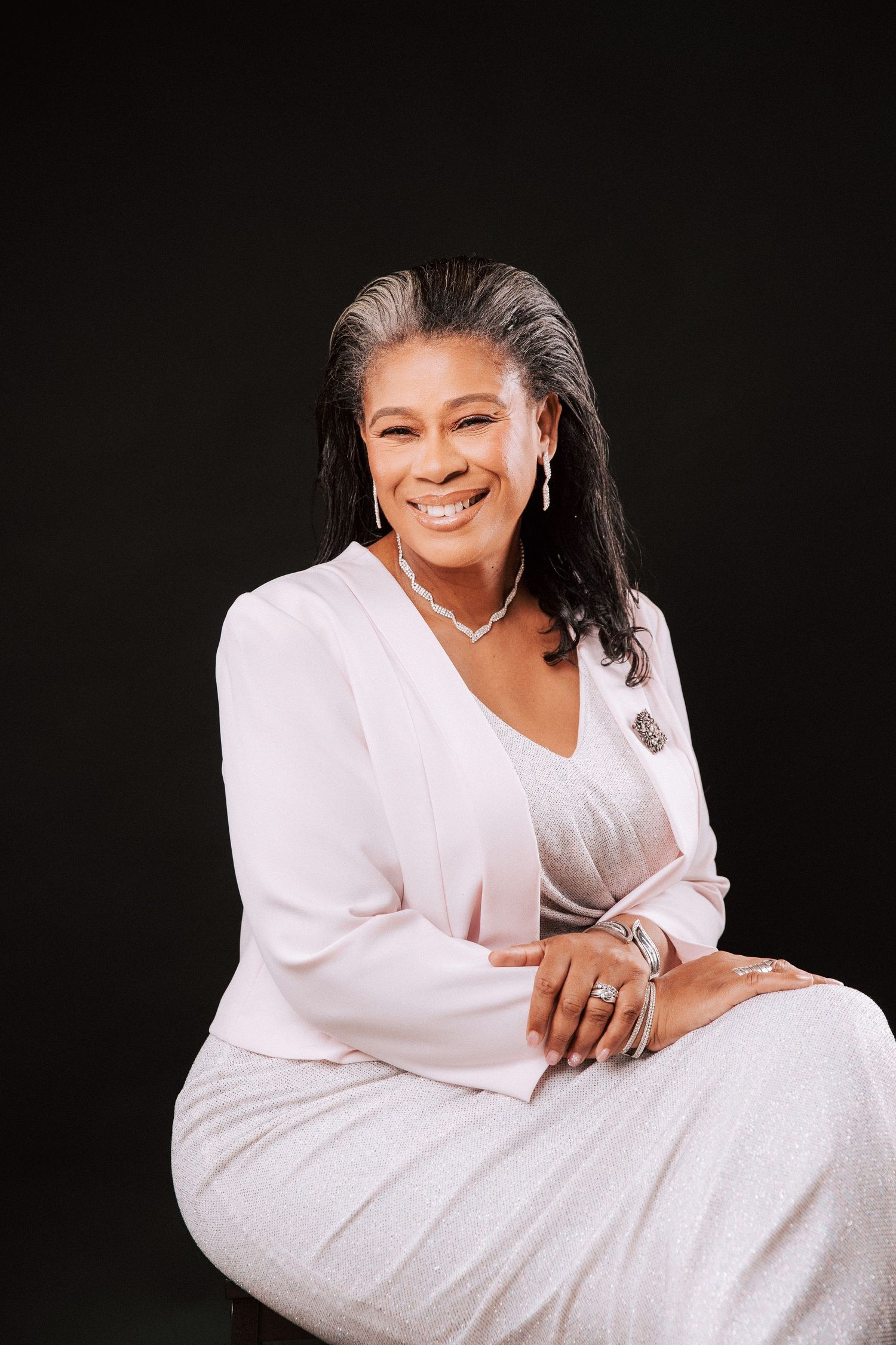 A woman sits smiling against a black background, wearing a pink blazer over a shimmery dress. an elegant choice for professional headshot