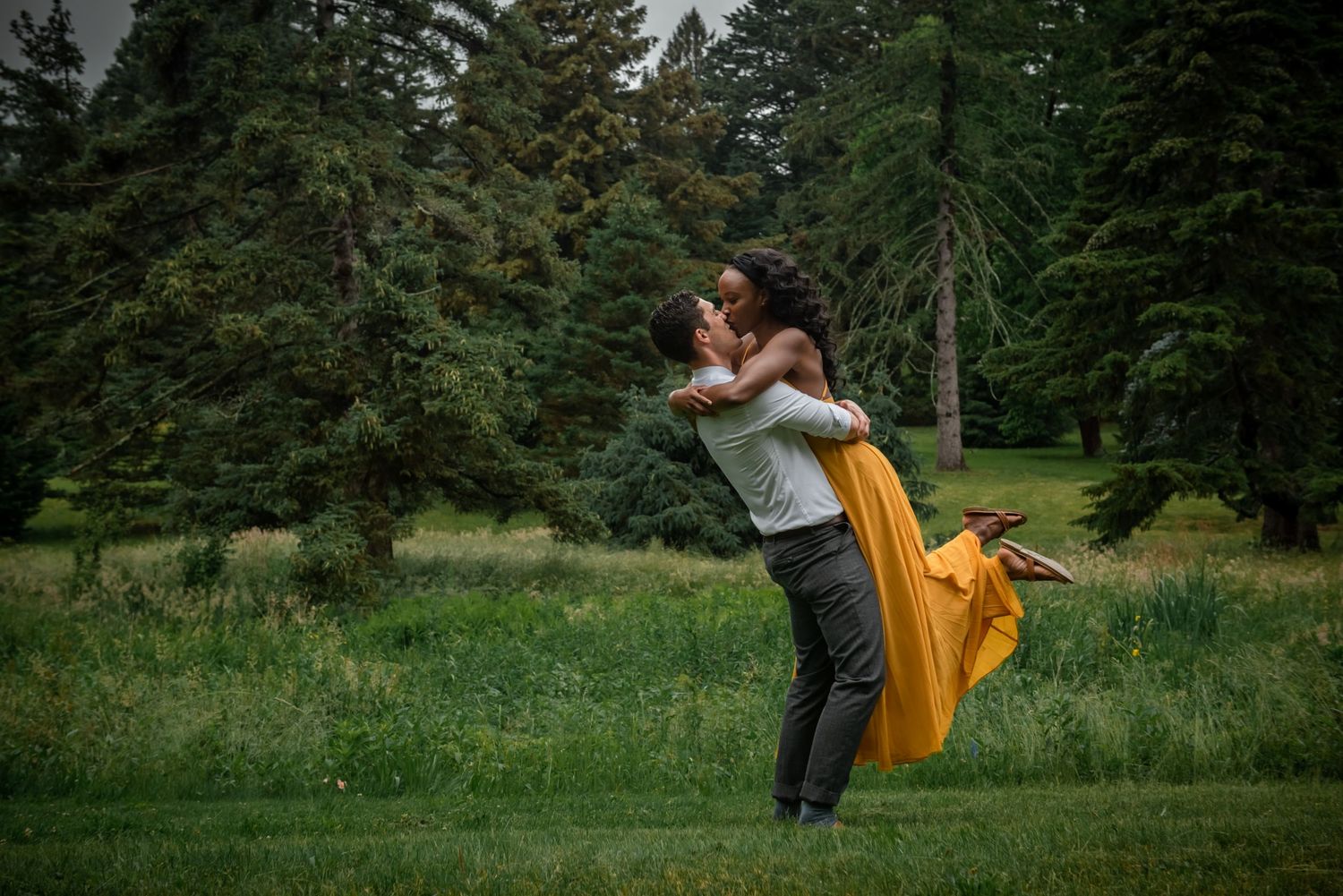 A couple embraces in a passionate pose amidst a lush green forest setting, with a flowing yellow dress adding vibrant color.