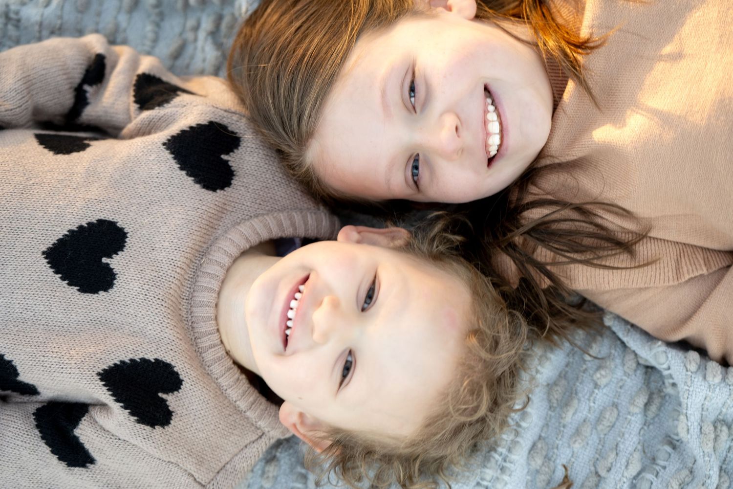 Two people wearing heart patterned sweaters lie head to head smiling on a gray surface.