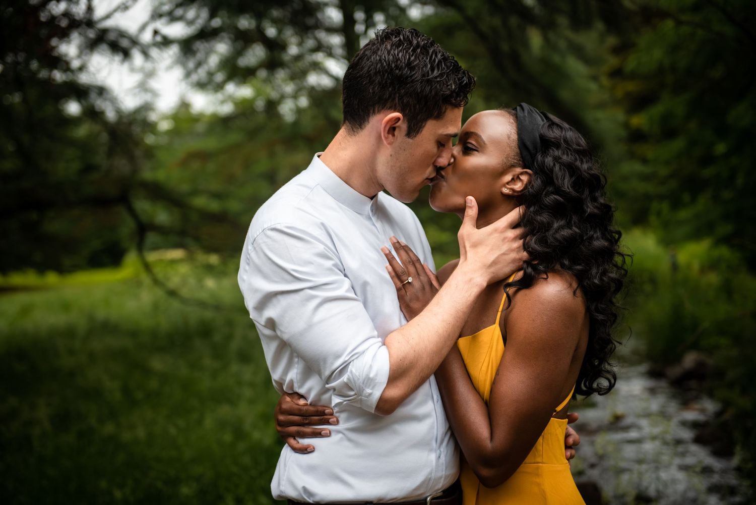 A romantic couple embraces and kisses in a lush green garden setting while wearing white and yellow outfits.