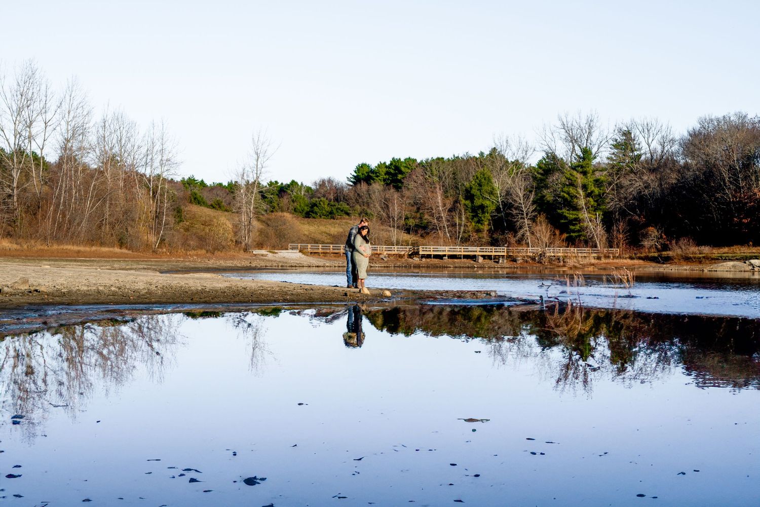 A figure stands on a frozen pond in winter with bare trees reflected in the icy surface during a clear day.