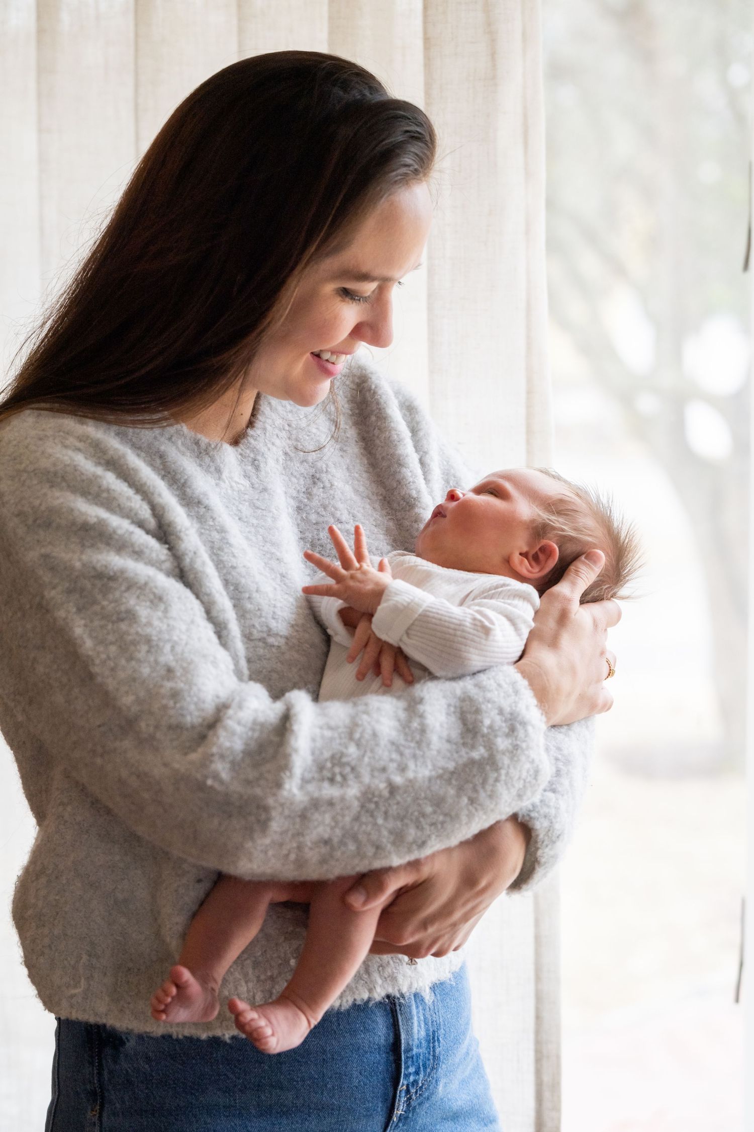 A tender moment between a mother in a cozy gray sweater holding her sleepy newborn baby by a bright window.