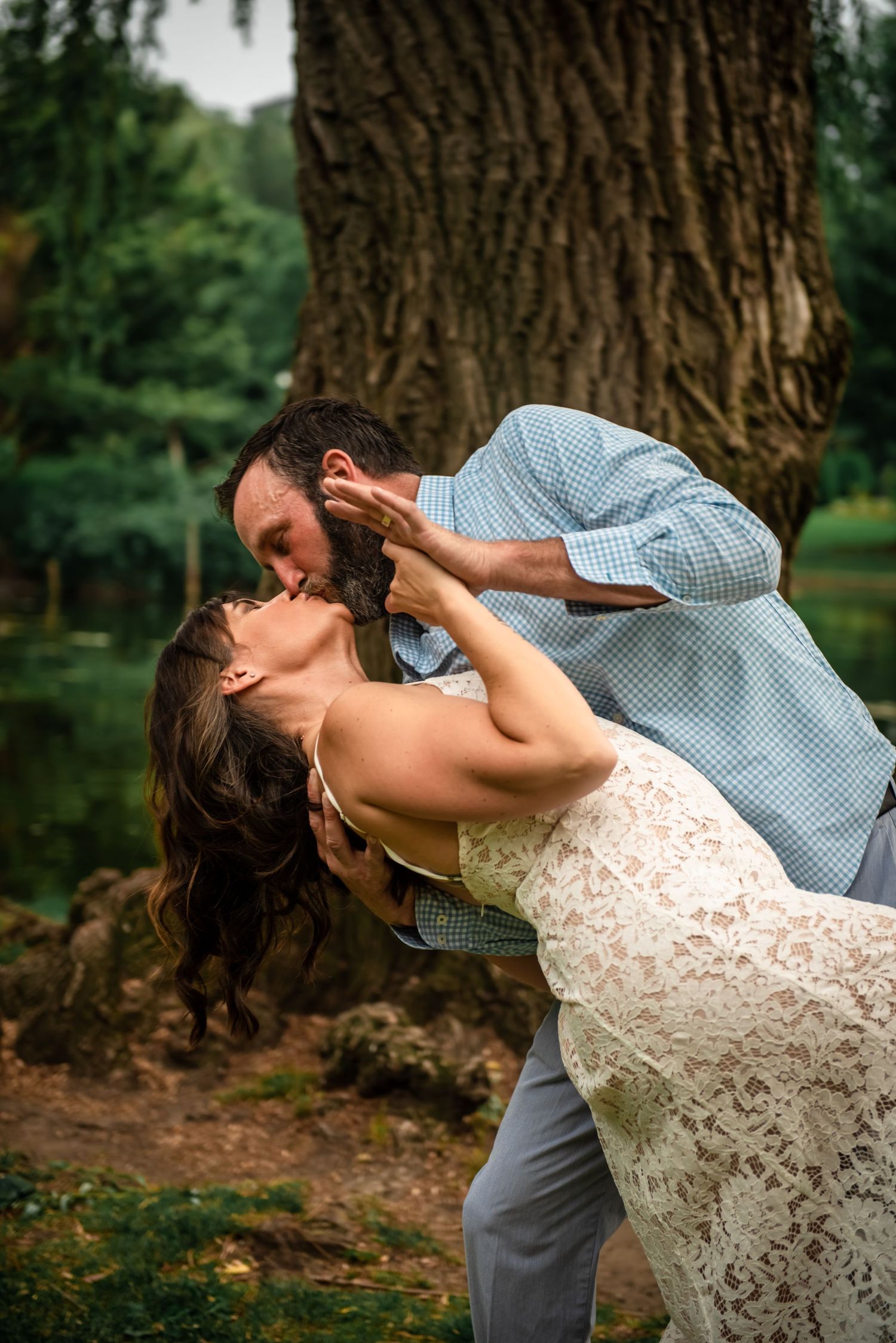 A romantic couple in wedding attire share an intimate dip kiss in front of a large tree trunk in a park setting.