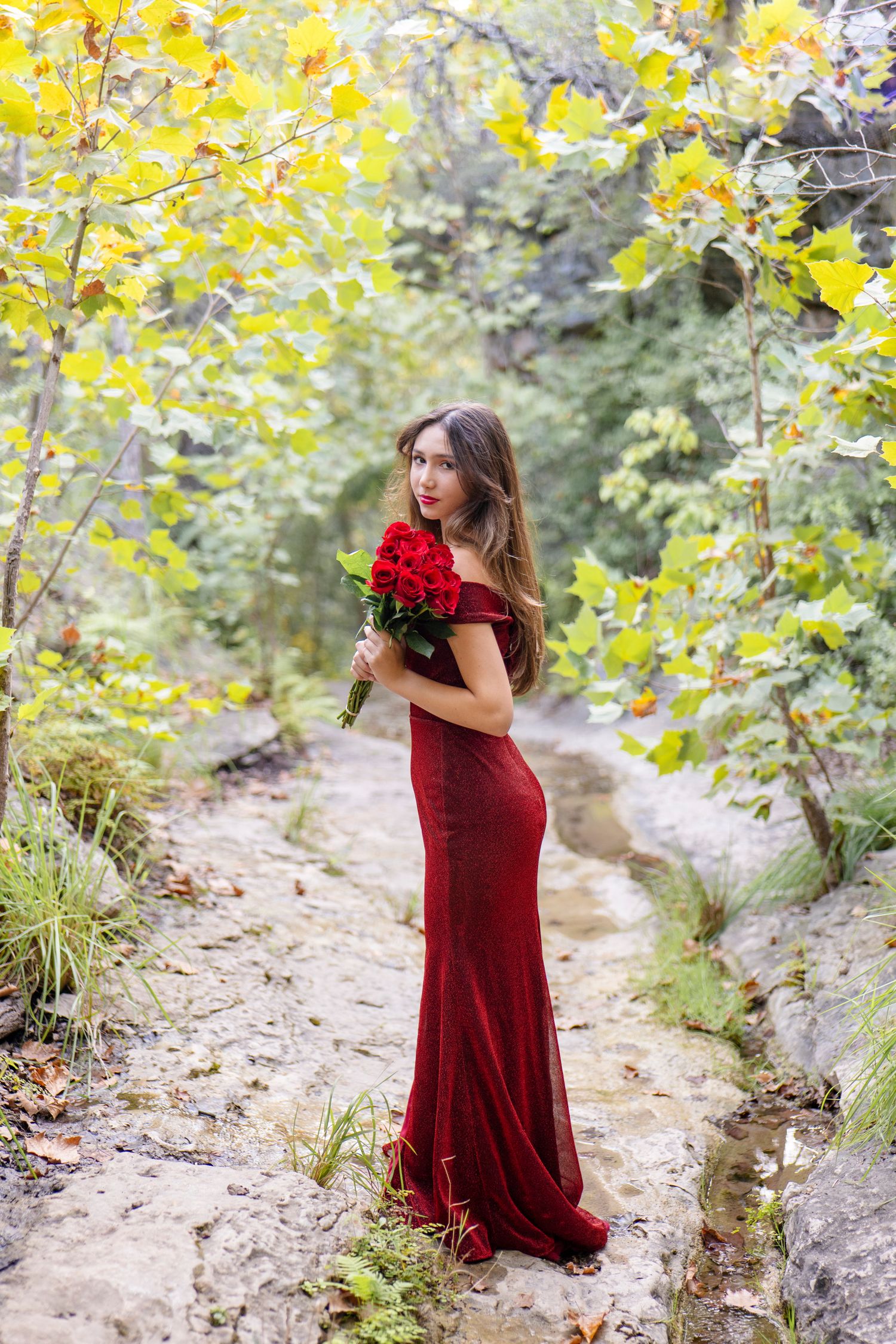 A person in a long burgundy formal gown holds red roses while standing on a nature path surrounded by yellow autumn leaves.