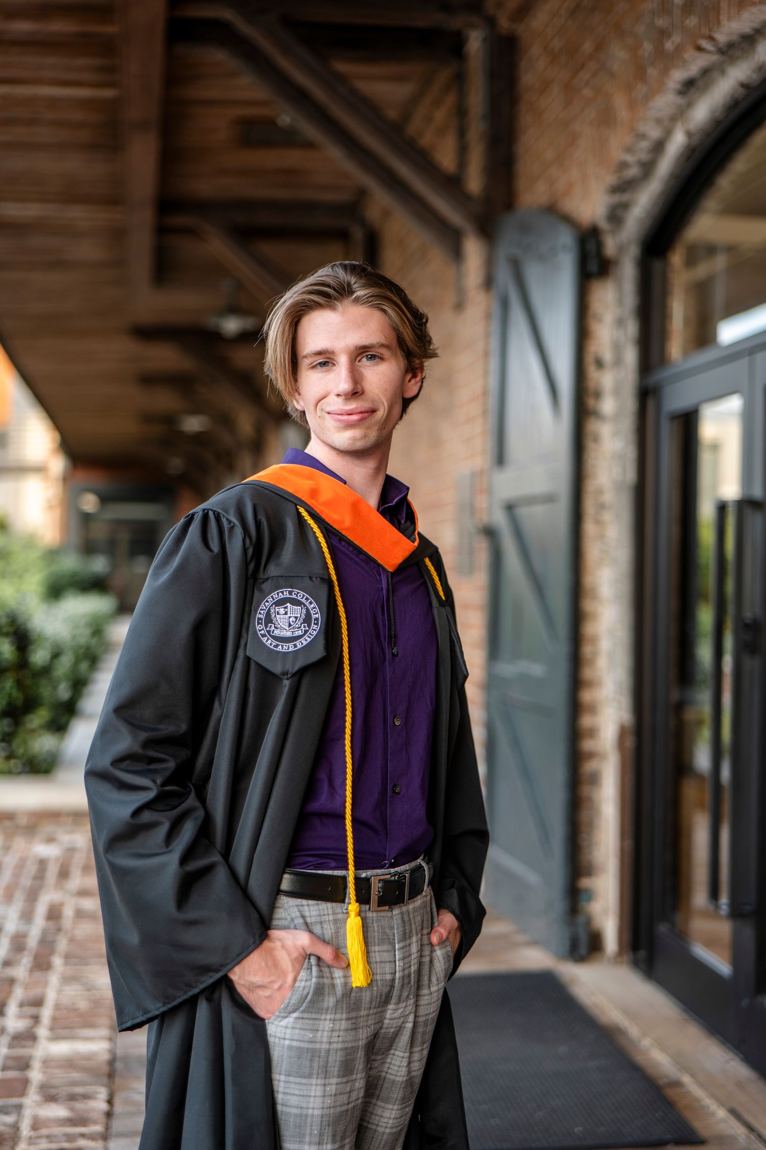 Graduate wearing academic regalia with orange stole and black gown poses in front of historic brick archway.