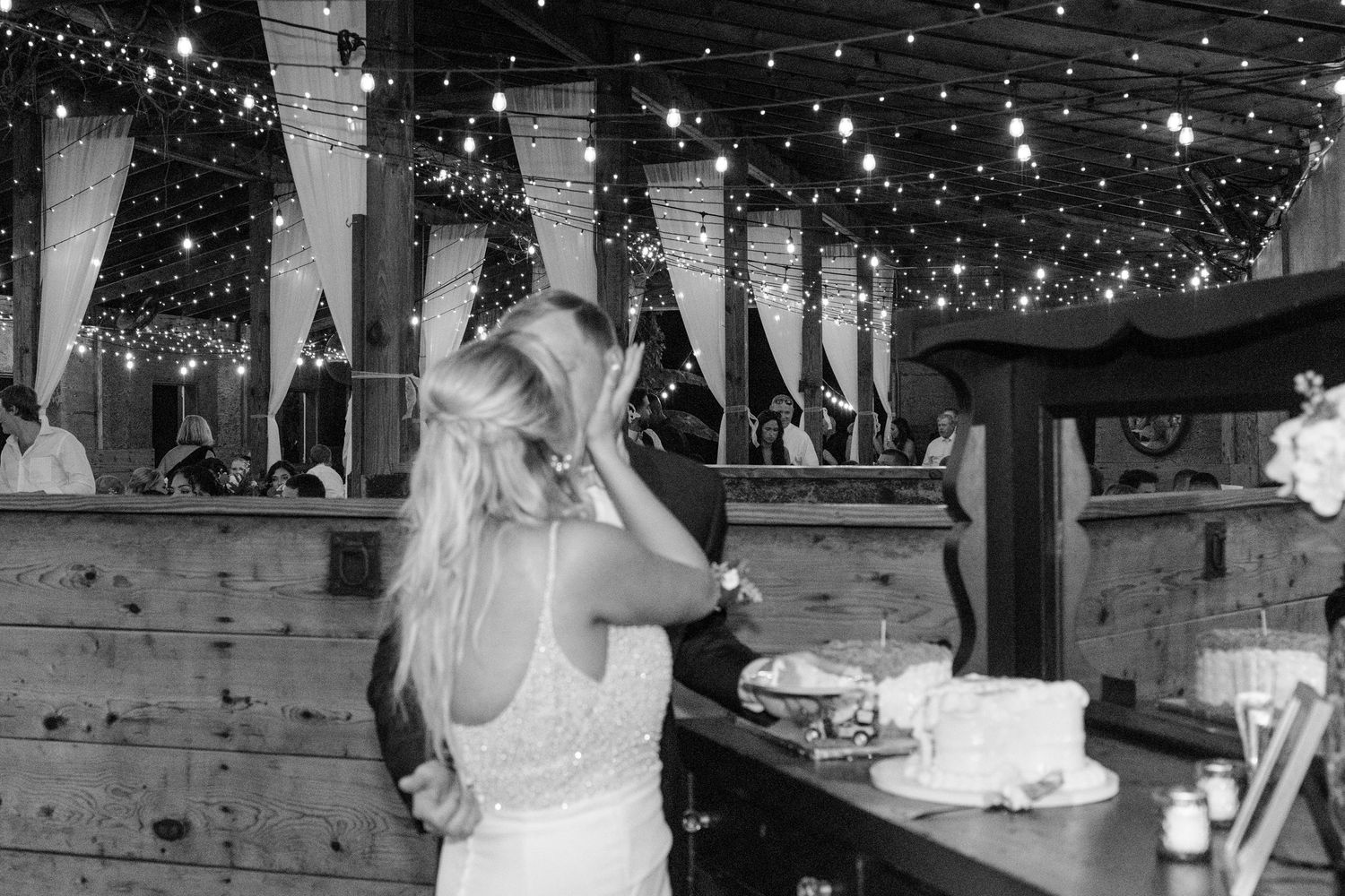 A bride enjoys her reception under twinkling string lights at a rustic wooden bar venue in black and white.